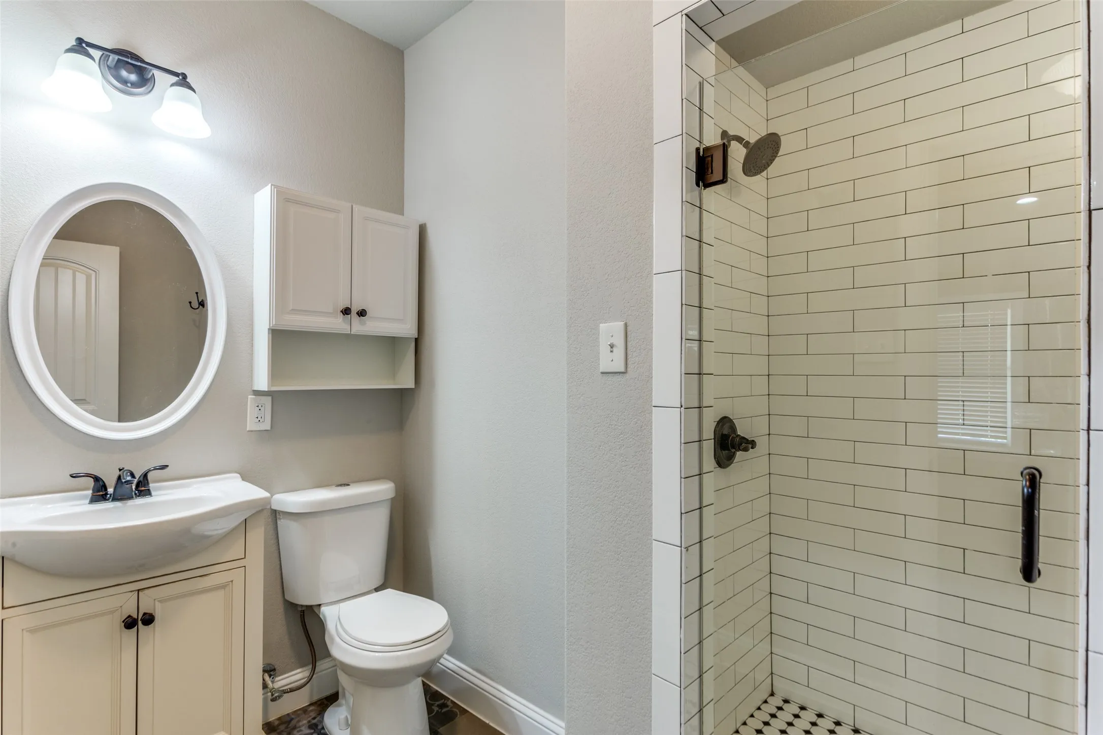 White oval-framed mirror, vanity with integrated basin, wall-mounted storage cabinet, glass-enclosed shower with subway tile surround, and hexagonal mosaic shower floor