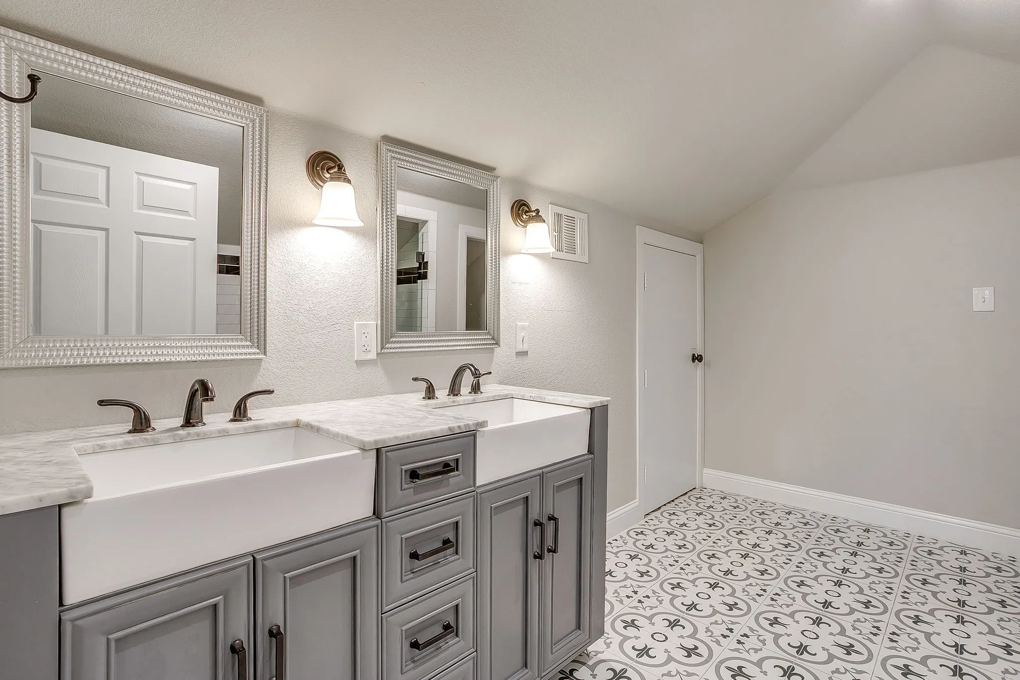 Dual vanity with farmhouse sinks, marble-finish countertops, gray cabinetry with dark hardware, and patterned floor tiling