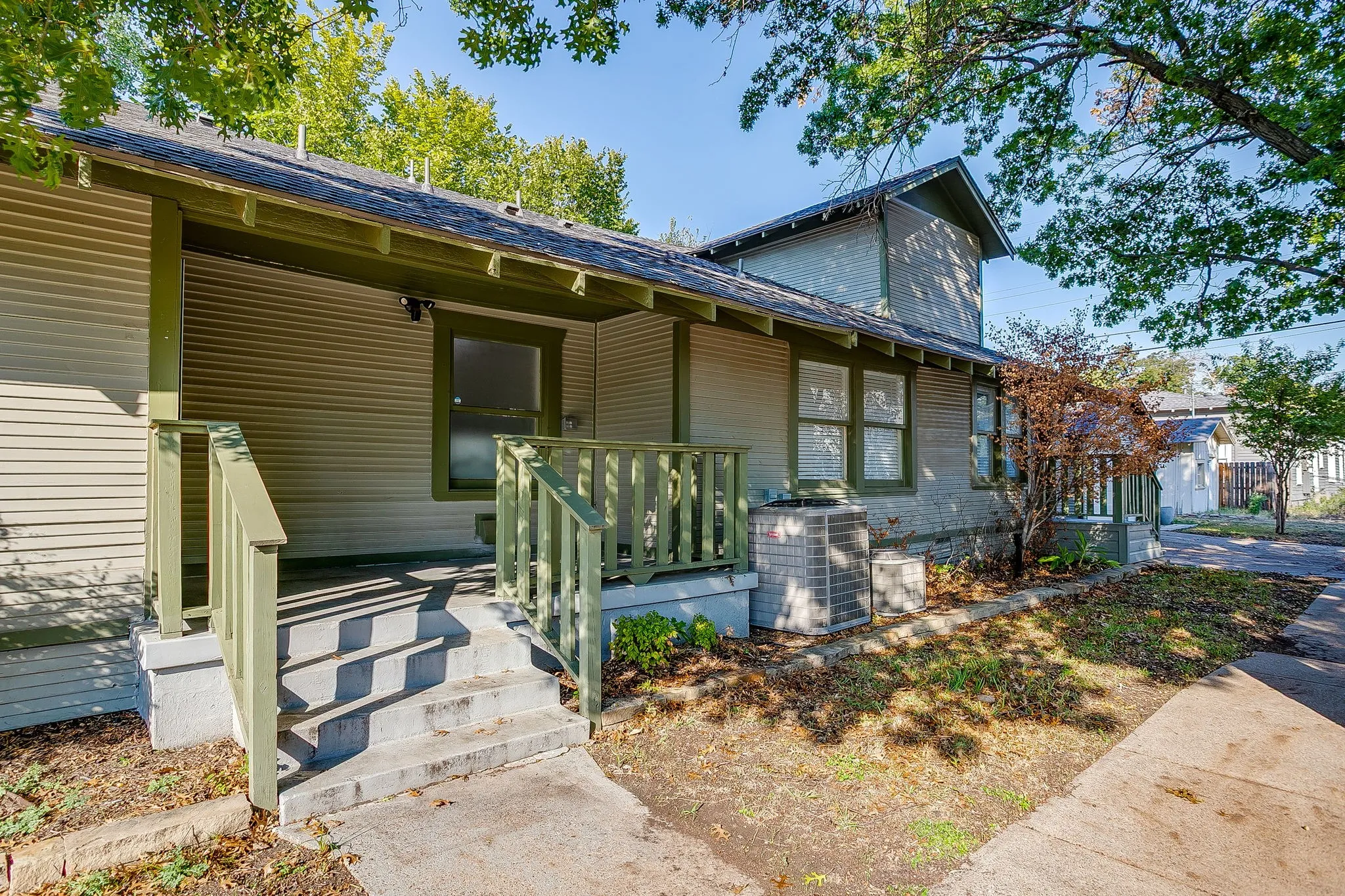 Covered porch with wood railing and concrete steps, horizontal siding exterior, multiple windows, and mature trees providing shade