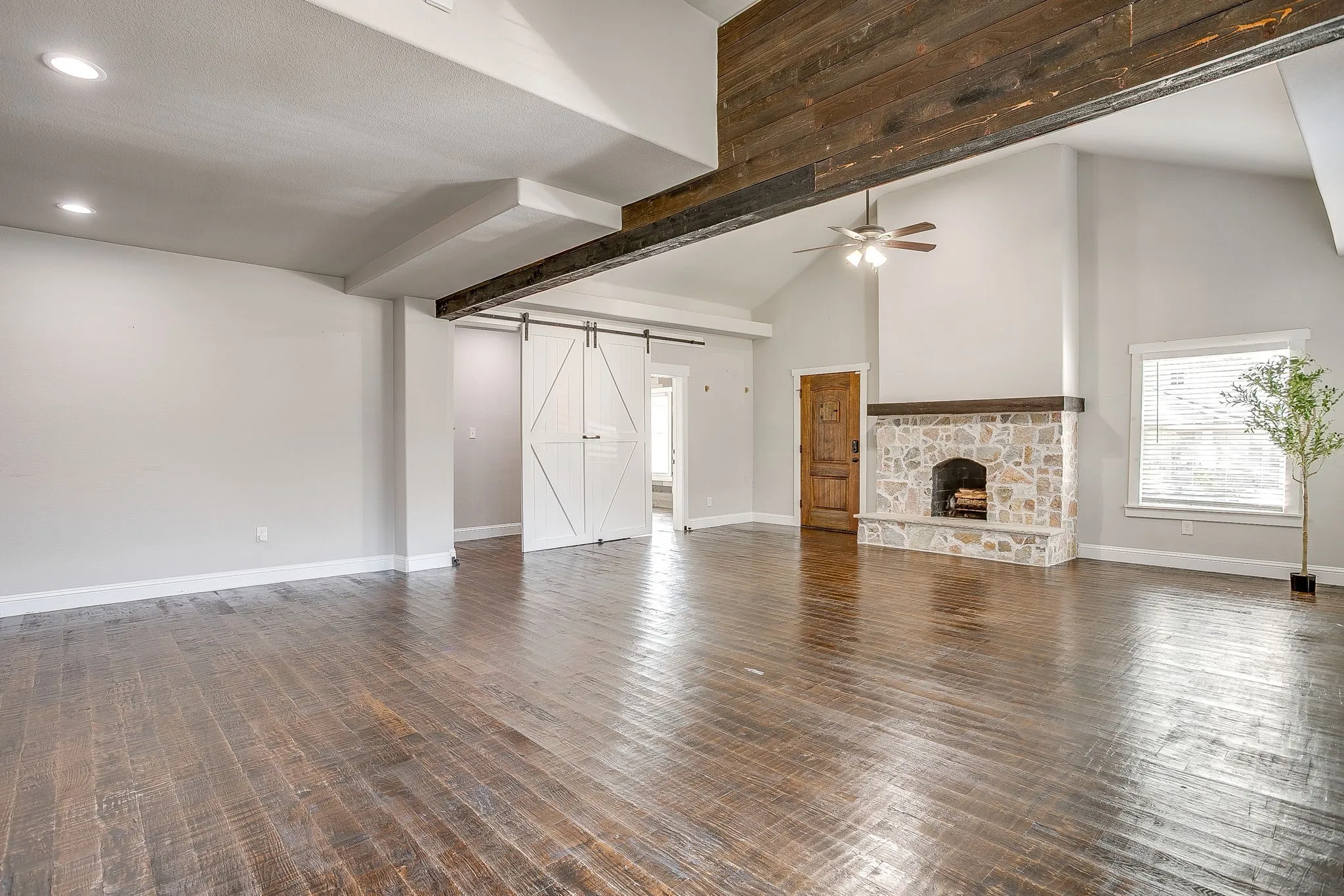 Vaulted ceiling with exposed wood beams, wood-finish flooring, stone-clad fireplace with mantel, ceiling fan, and white barn doors