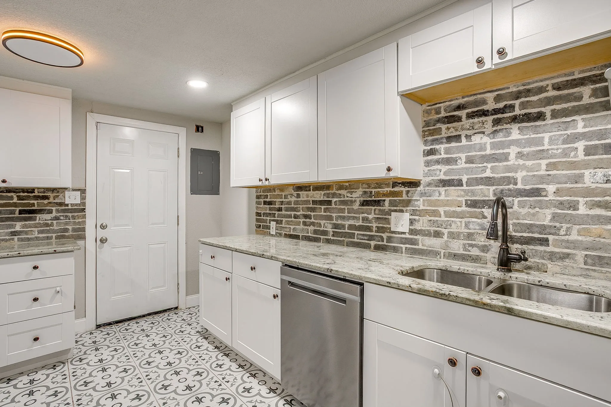 White shaker cabinetry, stainless steel dishwasher, dual basin sink with black gooseneck faucet, light-toned stone countertop, and brick-patterned backsplash