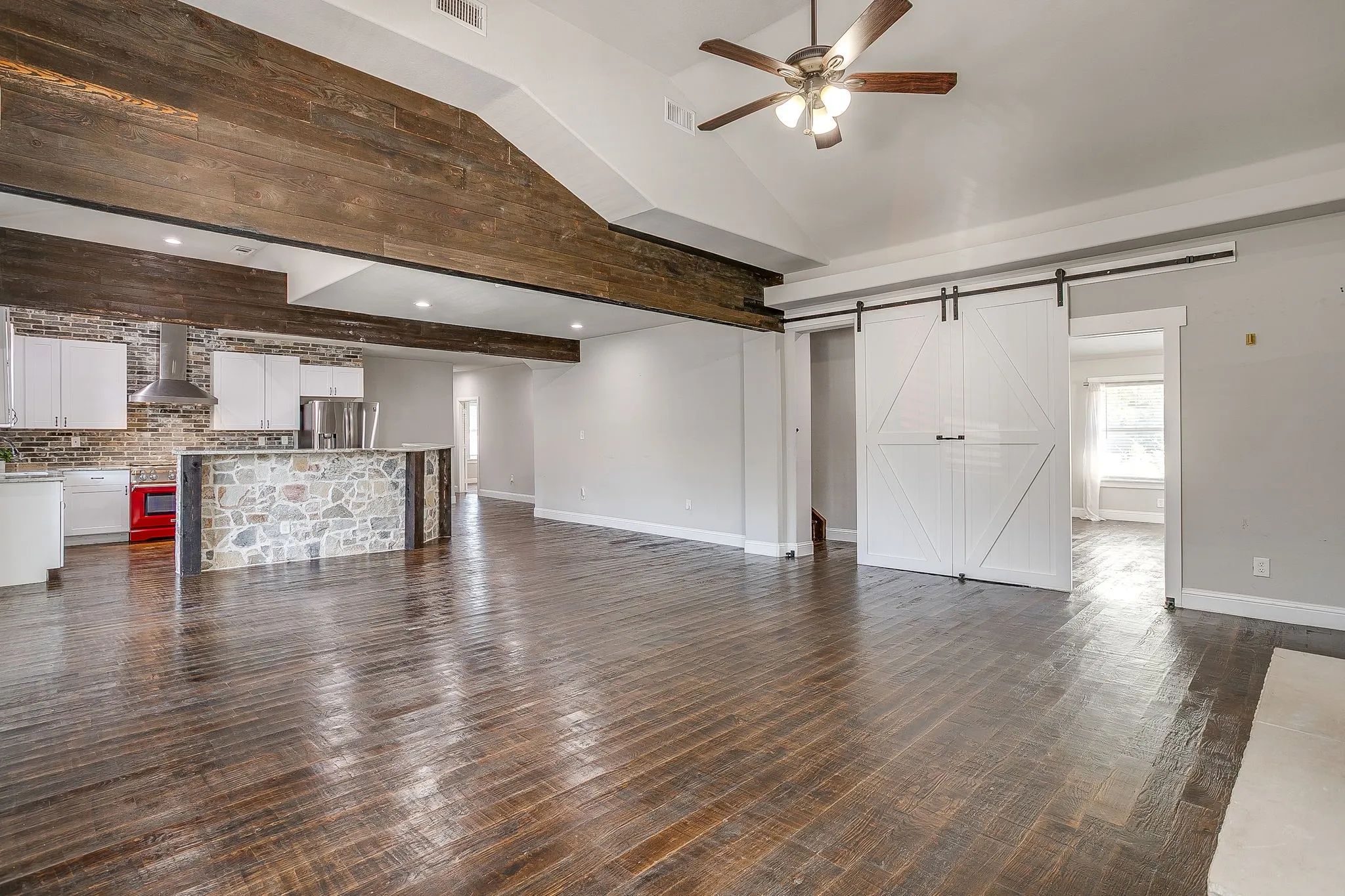Vaulted ceiling with wood-finish beams, exposed brick kitchen wall, stone-front island with white cabinetry, barn doors, and dark wood-finish flooring