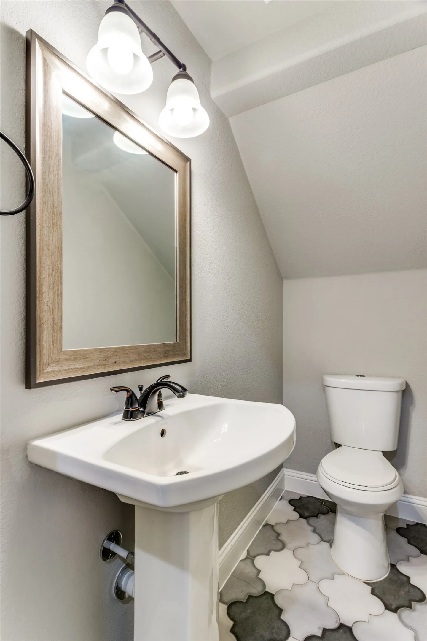 Pedestal sink, wall-mounted light fixture, decorative framed mirror, two-tone patterned floor tile, and white toilet
