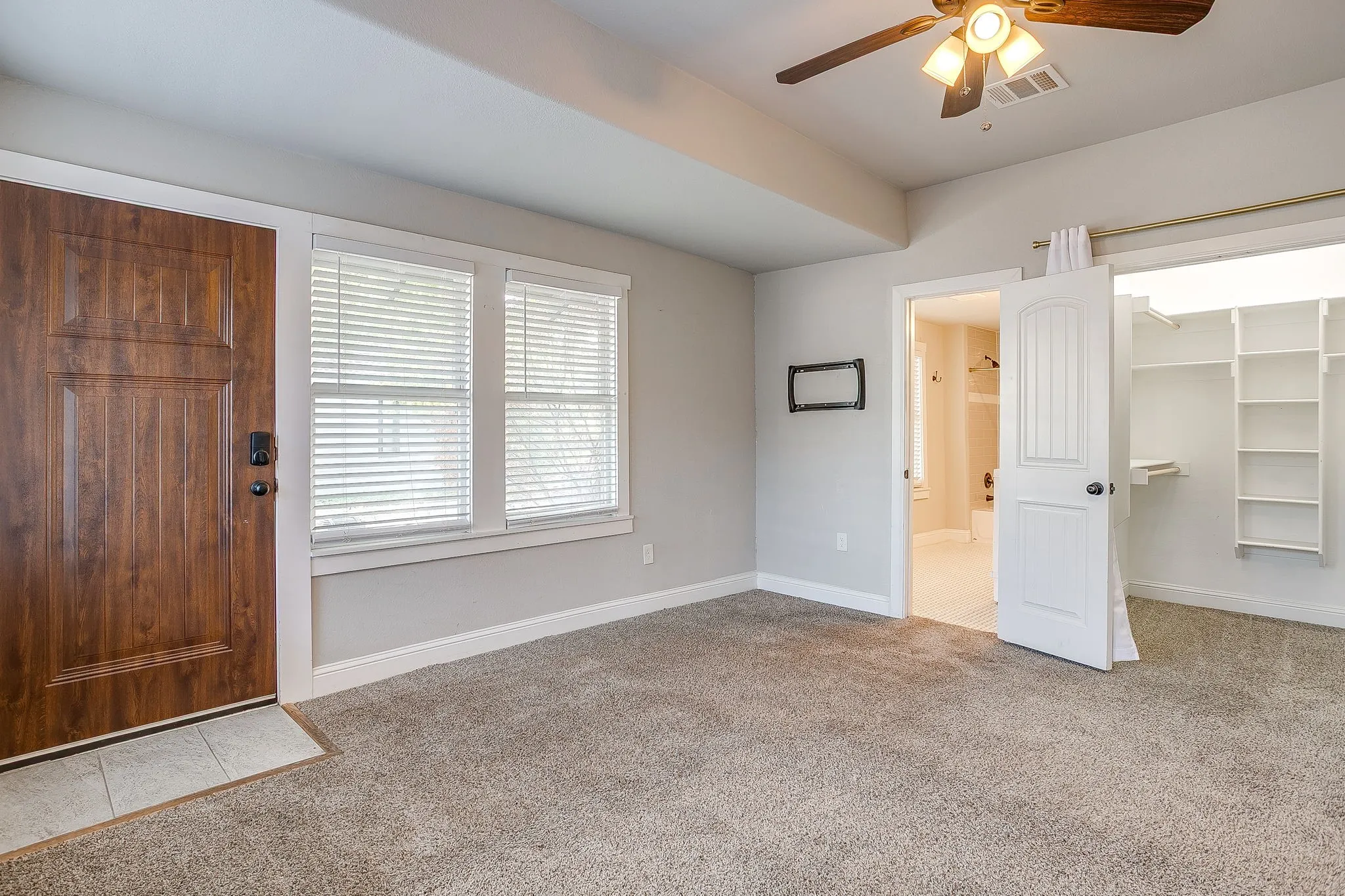 Wood-finish entry door, two windows with blinds, ceiling fan with integrated lighting, built-in closet shelving, and a tiled floor visible through the doorway