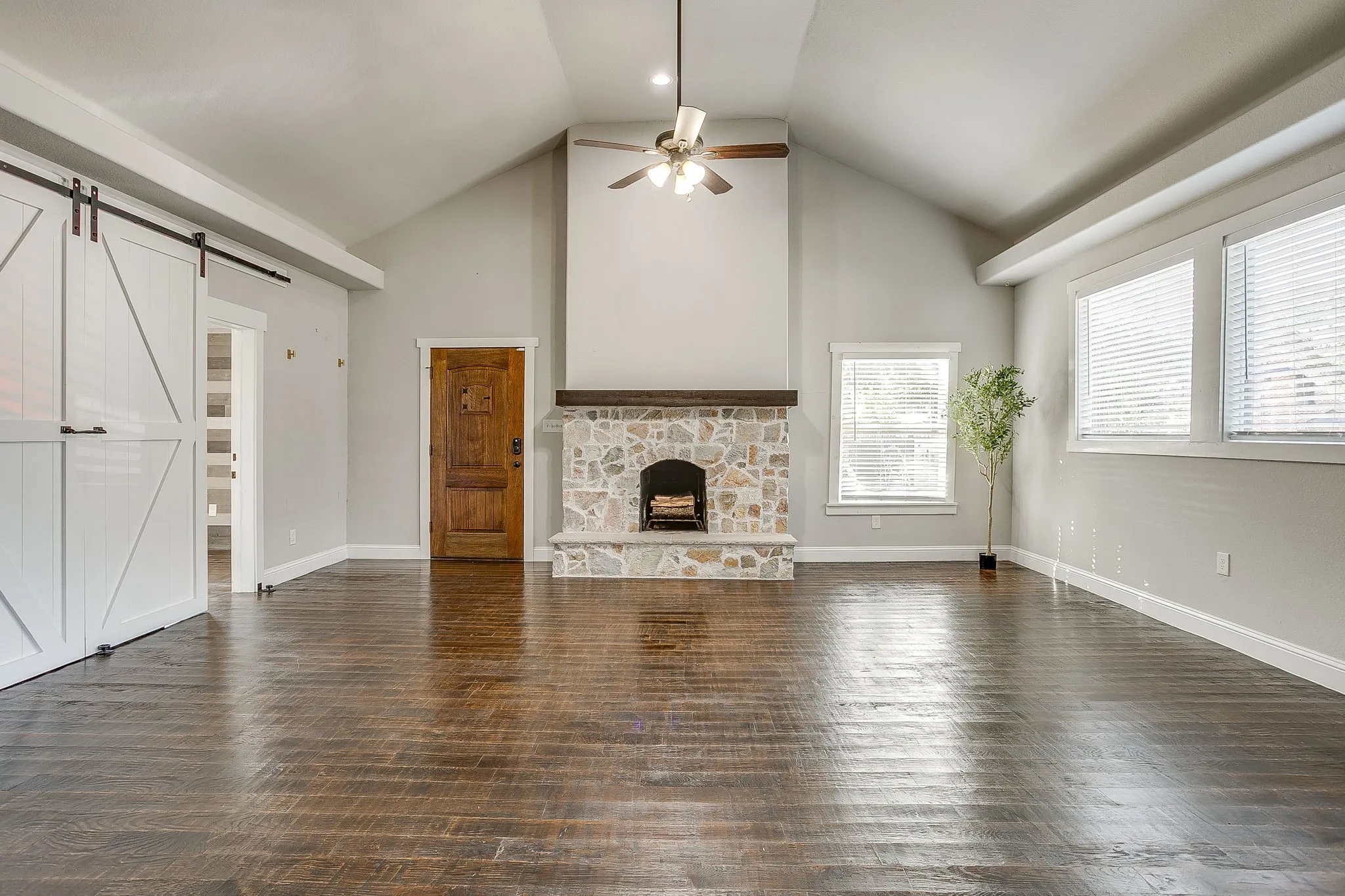 Vaulted ceiling with a ceiling fan, stone-clad fireplace with wood mantle, wood-finish flooring, two large windows, and a barn door entry