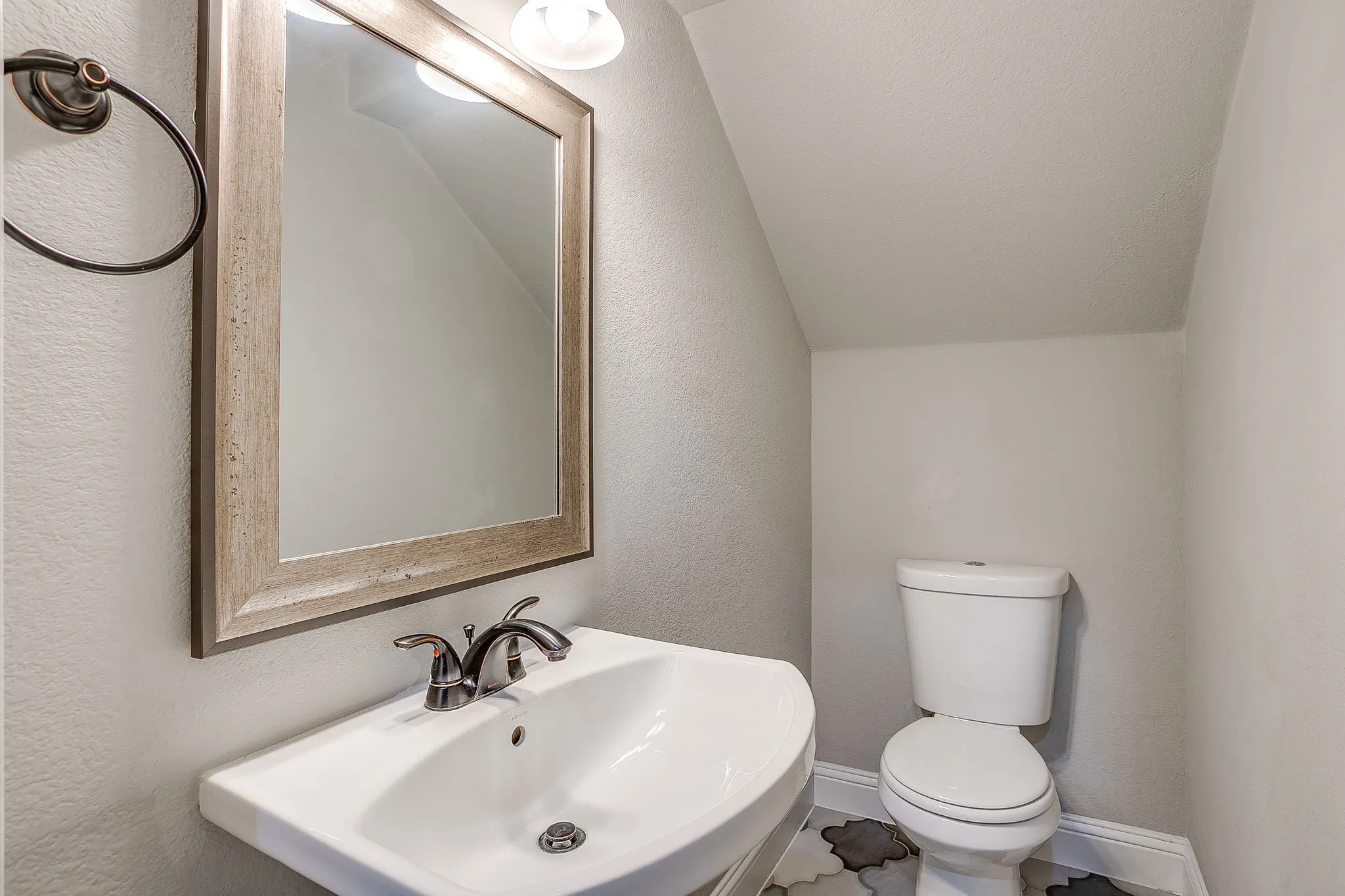 Pedestal sink with dark bronze faucet, framed mirror with vanity lighting, white toilet, and patterned floor tile