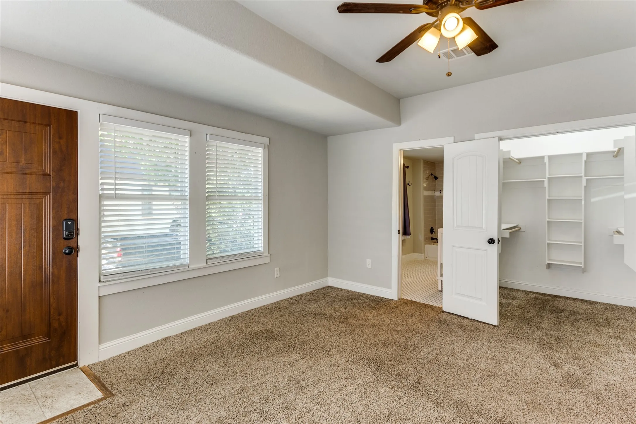 Wood-finish entry door, two large windows with blinds, ceiling fan with integrated lighting, custom closet shelving, and textured carpeting