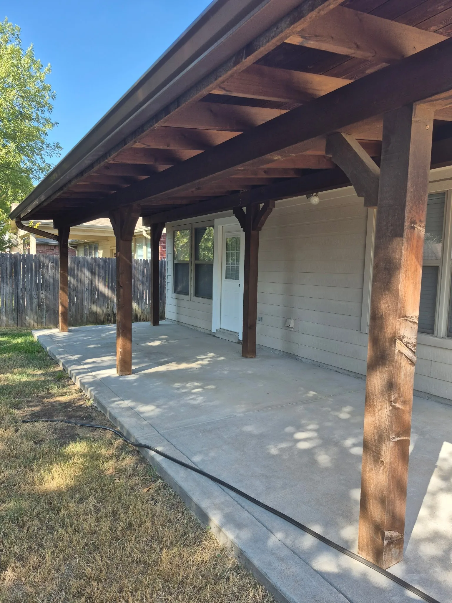 Covered concrete patio with exposed wood beam ceiling, wood support columns, and adjacent lawn area