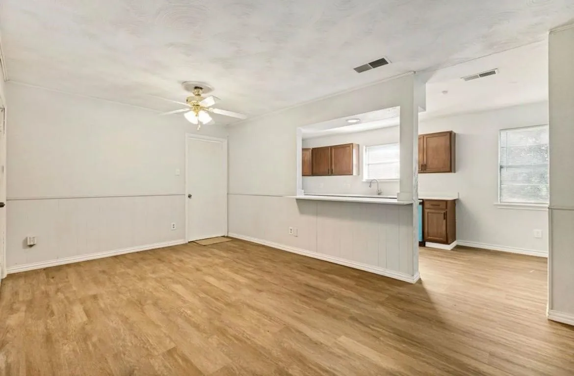 Wood-finish flooring throughout, white paneled wainscoting, ceiling fan with light fixture, and an open pass-through to the kitchen featuring wood cabinetry