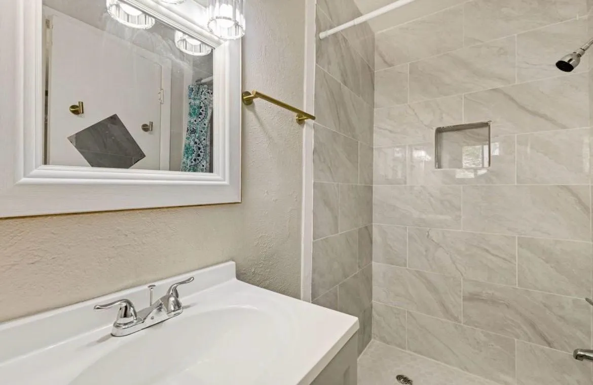 Bathroom featuring a vanity with a white countertop, chrome faucet, framed mirror, and a shower with light-toned tiled walls and a recessed niche
