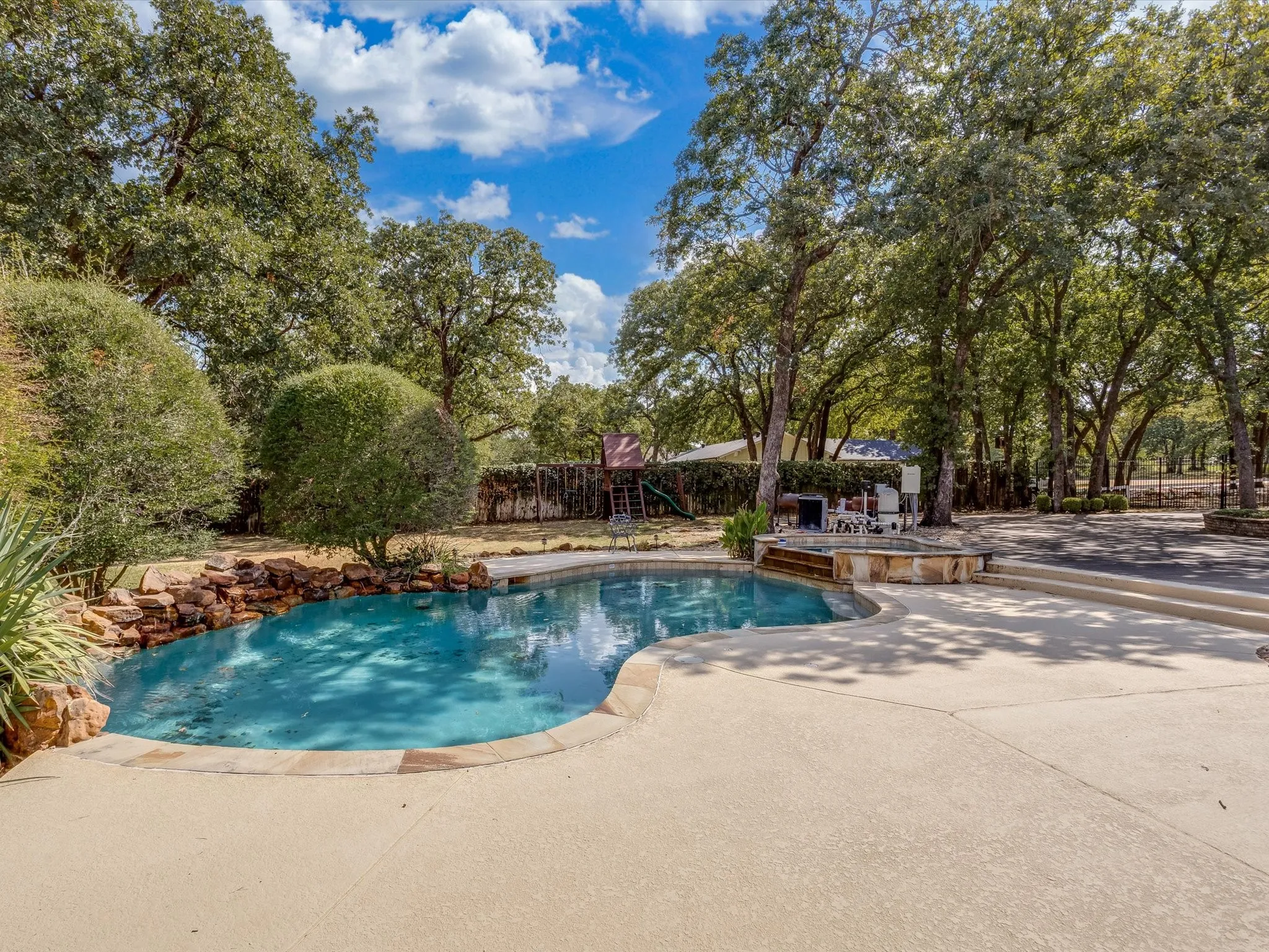 Curved swimming pool with a rock waterfall feature, integrated spa, and expansive concrete decking, surrounded by mature trees