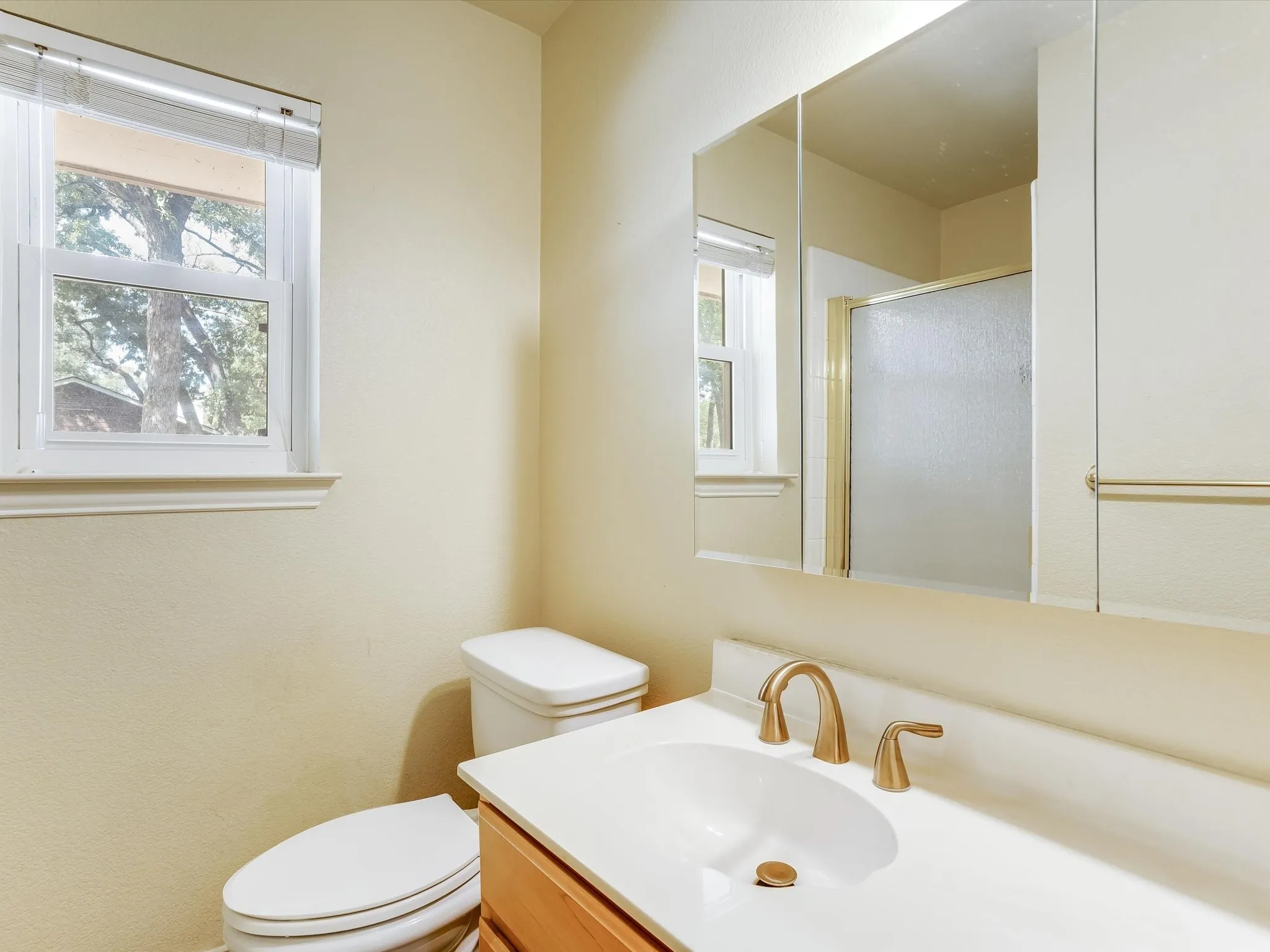 White single-pane window, white vanity with integrated sink, gold-toned faucet, recessed mirrored medicine cabinet, and shower enclosure with frosted glass