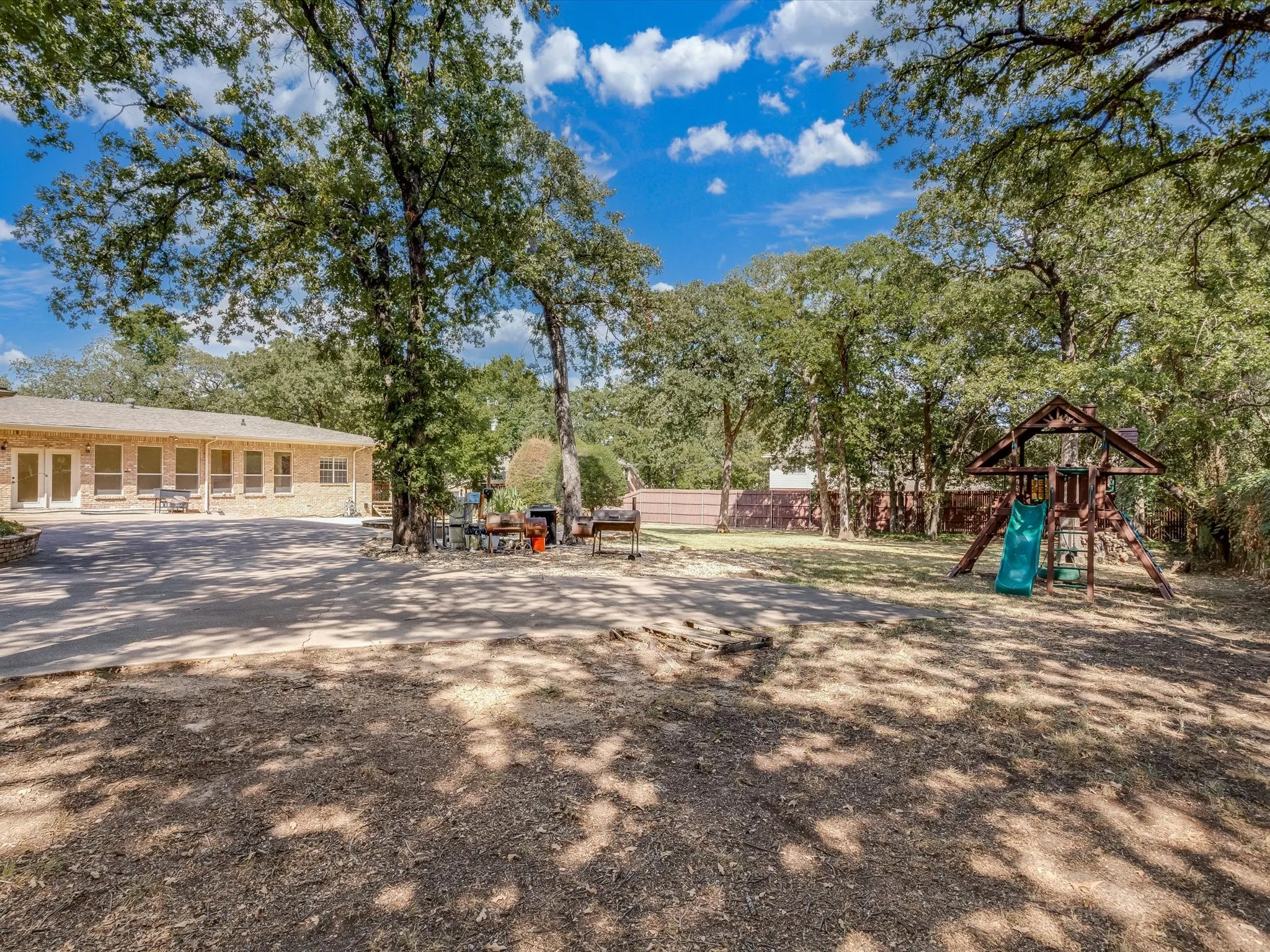 Expansive outdoor area featuring mature trees, a brick-clad structure with multiple windows and doors, and a spacious paved surface