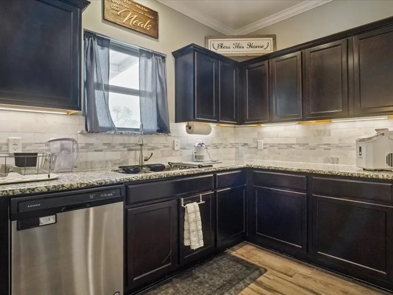 Dark wood-finish cabinetry, granite-style countertops, stainless steel dishwasher, tile backsplash, and under-cabinet lighting