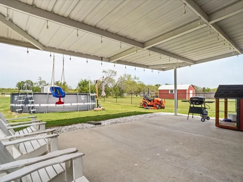 Covered concrete patio with string lighting, overlooking a green lawn with a large above-ground pool, and a red barn structure