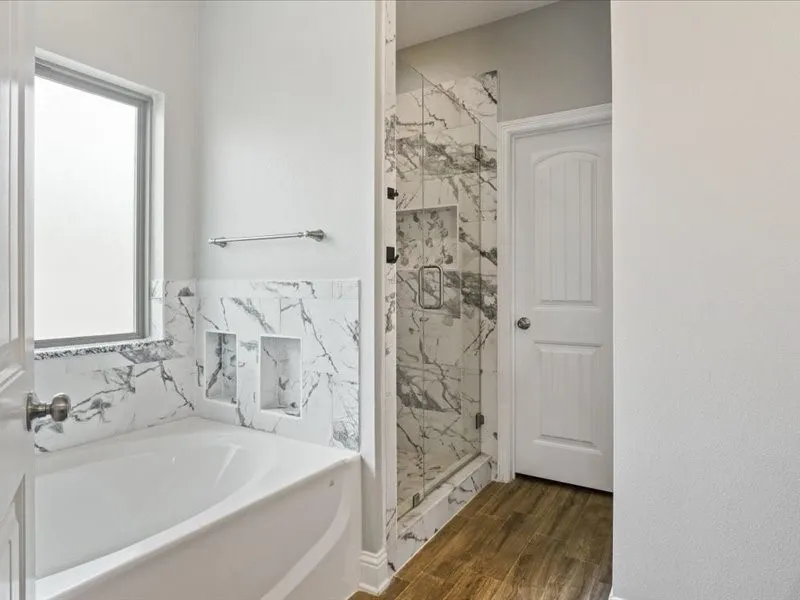 Bathtub with surrounding marble-look tile, glass-enclosed shower with tile surround, and wood-finish flooring