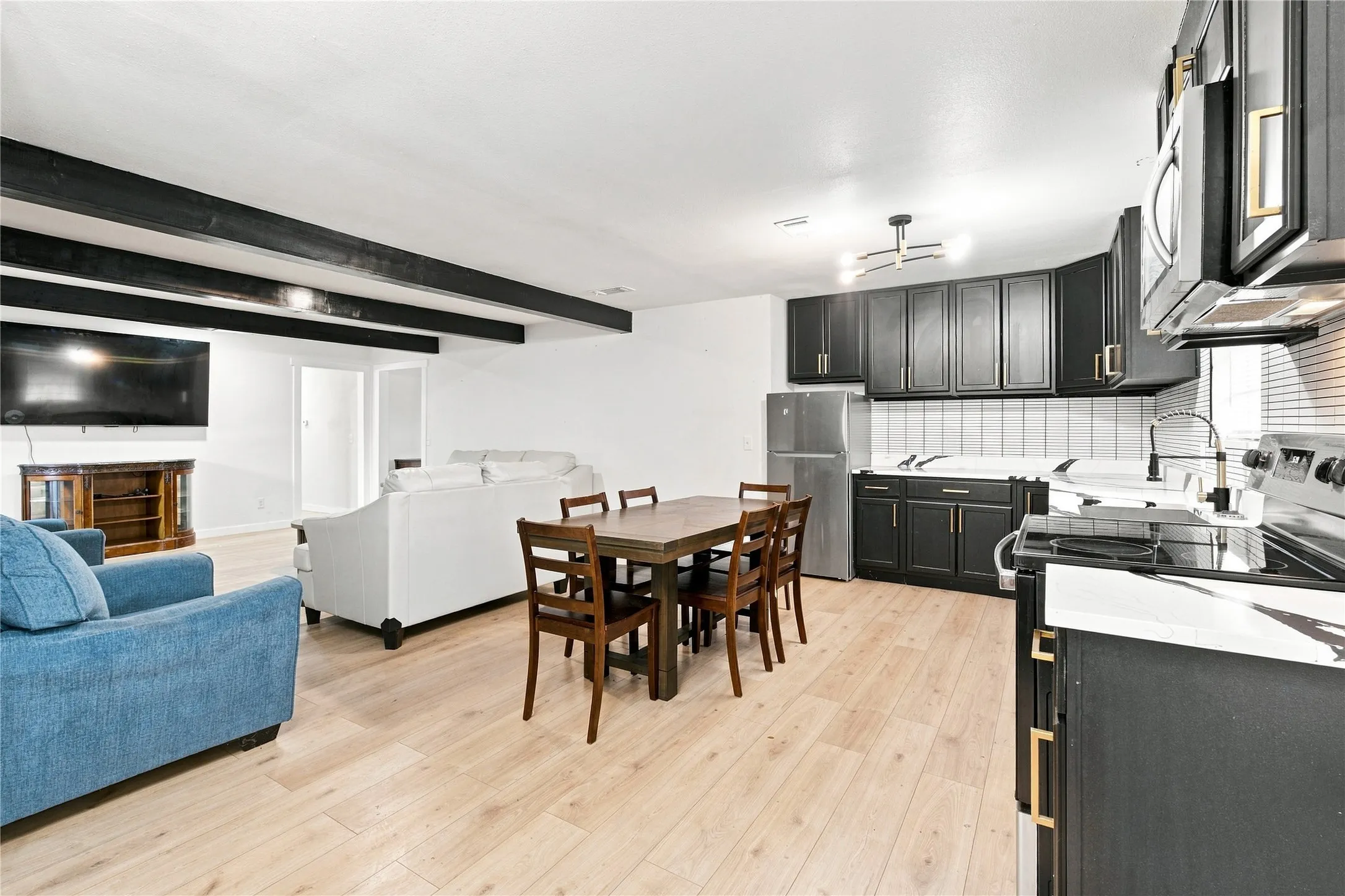 Open-concept living space with light wood-finish flooring, exposed ceiling beams, and a full kitchen featuring dark cabinetry, stainless steel appliances, and white countertops