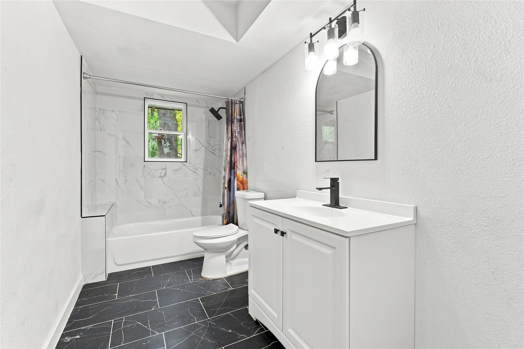 Bathroom featuring a white vanity with integrated sink, matte black faucet, arched mirror, contemporary lighting fixture, and a bathtub with white-veined tile surround