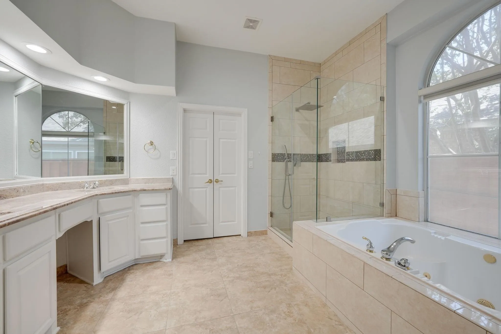 Dual vanity with integrated sinks, built-in soaking tub, glass-enclosed shower with tile surround, arched window, and tile flooring