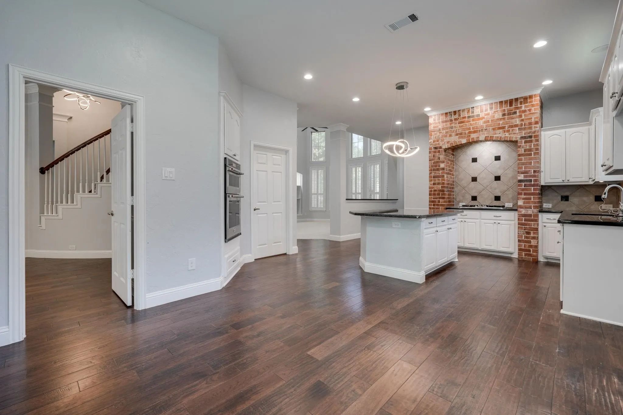 Wood-finish flooring, white cabinetry, dark countertops, brick accent wall, and a central island with pendant lighting