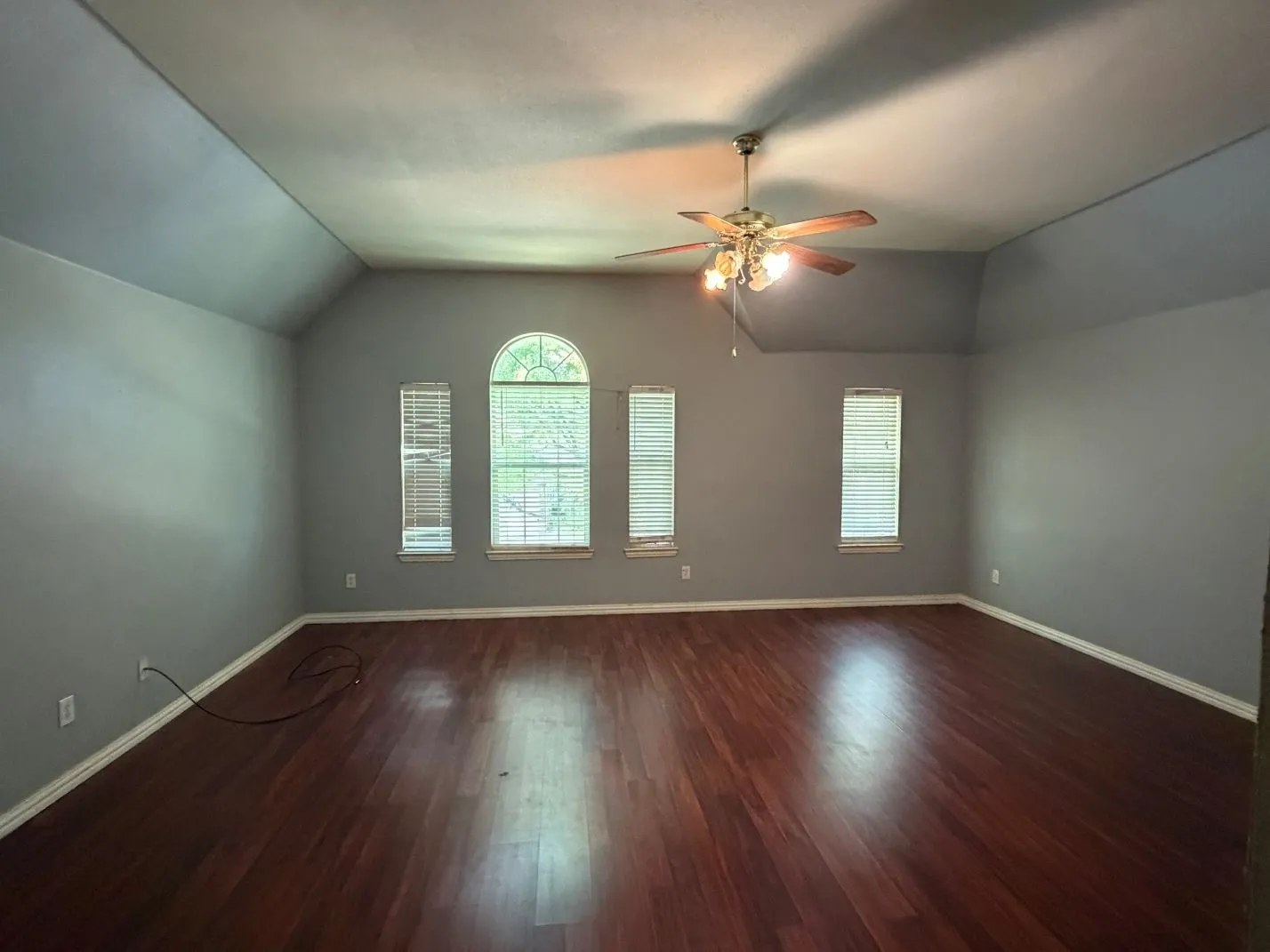 Wood-finish flooring, vaulted ceiling, arched window, ceiling fan with integrated lighting, and multiple windows with blinds