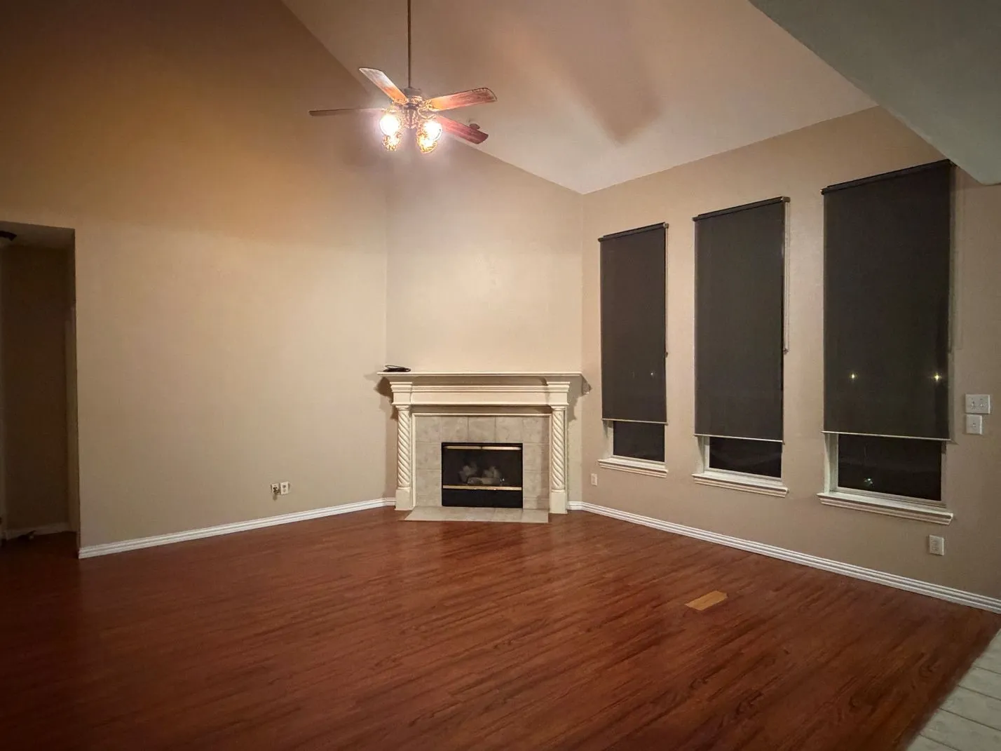 Vaulted ceiling with a ceiling fan, fireplace with a decorative mantel and tile surround, wood-finish flooring, and multiple windows with roller shades