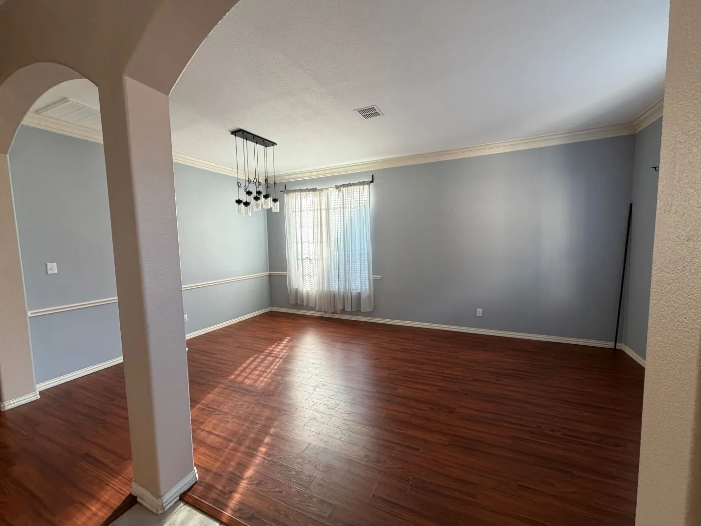 Expansive room with wood-finish flooring, an arched entryway, a window with sheer coverings, an updated chandelier, and crown molding