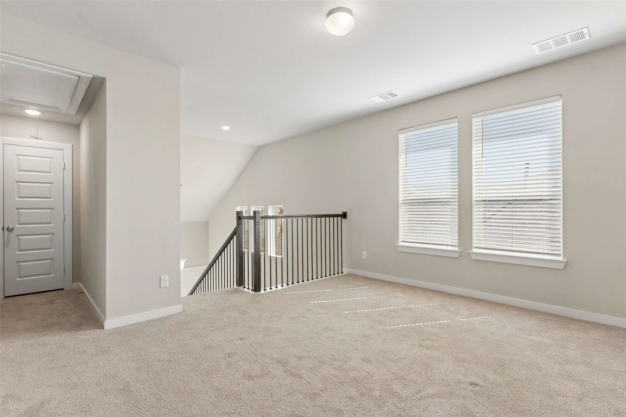 Carpeted room with a staircase and wood-finish railing, two windows with horizontal blinds, and a paneled interior door