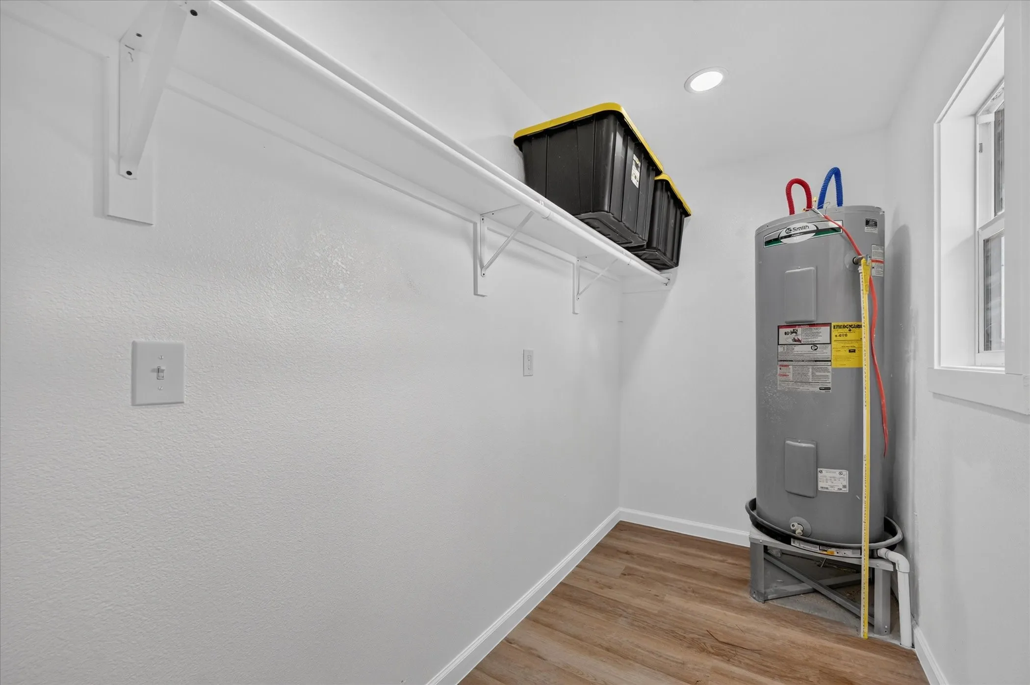 Utility area featuring wood-finish flooring, a water heater, a window, built-in shelving, and recessed lighting