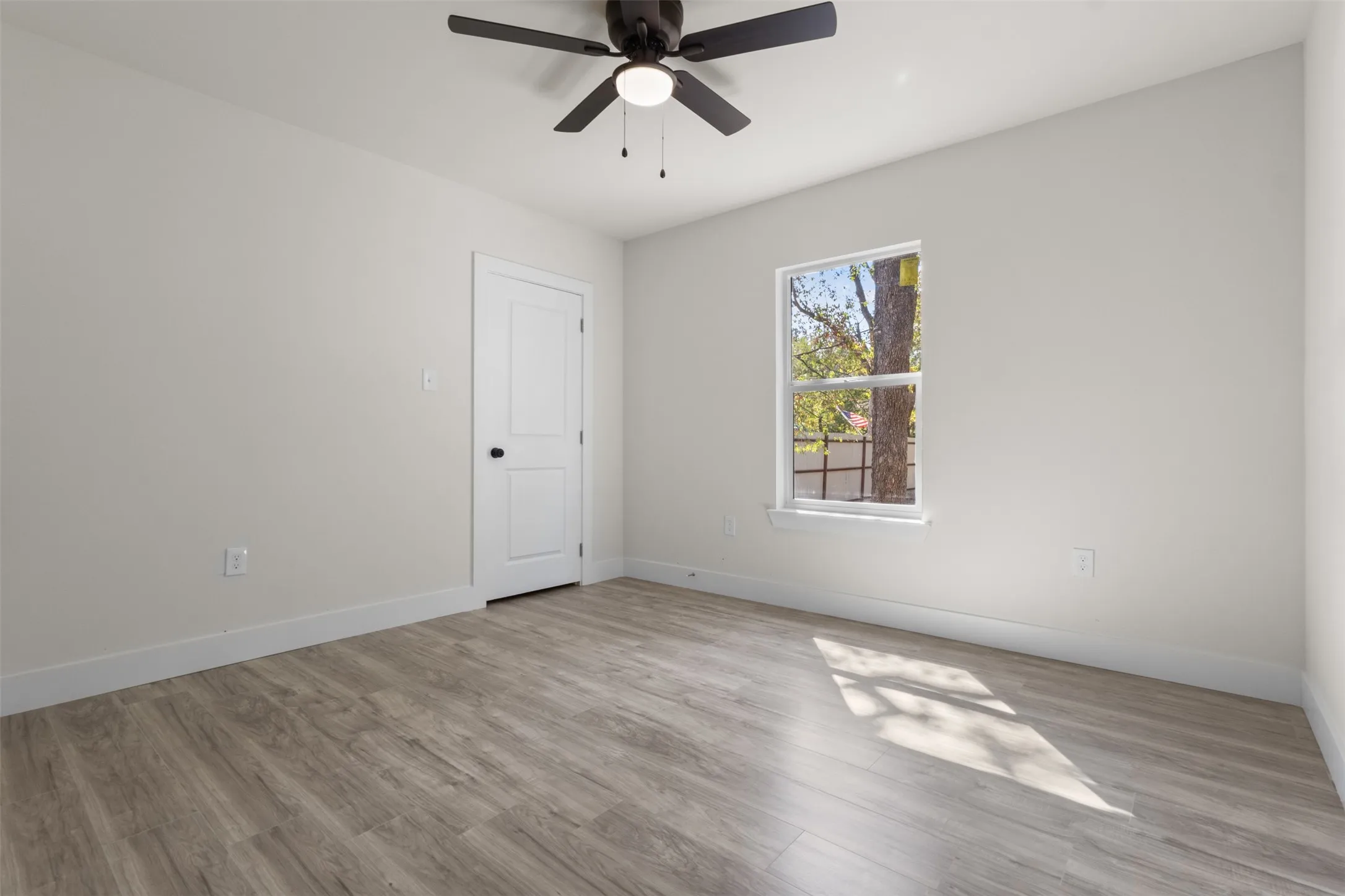 Spacious room featuring wood-finish flooring, a ceiling fan with integrated lighting, a two-panel interior door, and a single window with white trim
