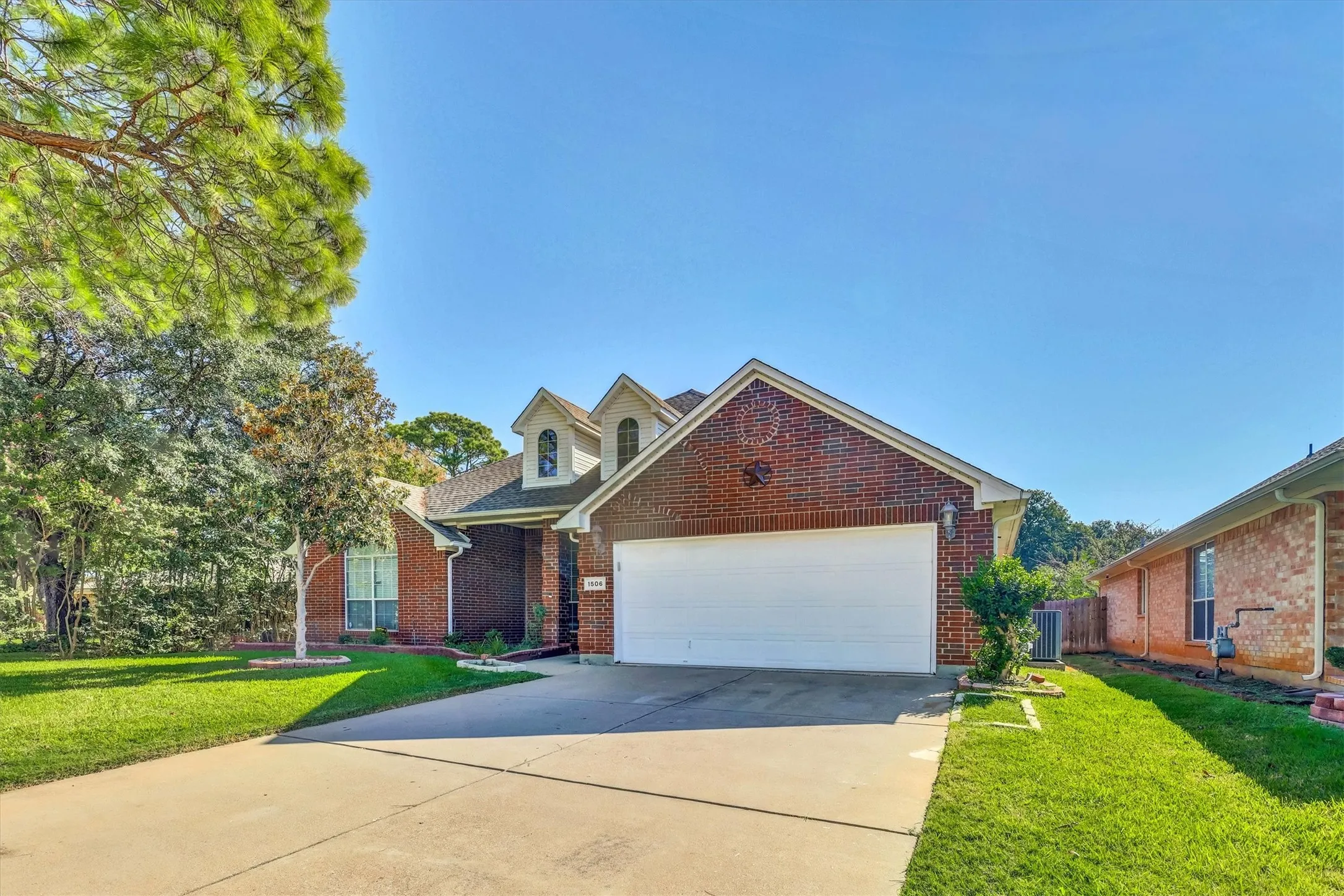 Brick exterior with a two-car garage, mature trees, a well-maintained lawn, and a concrete driveway