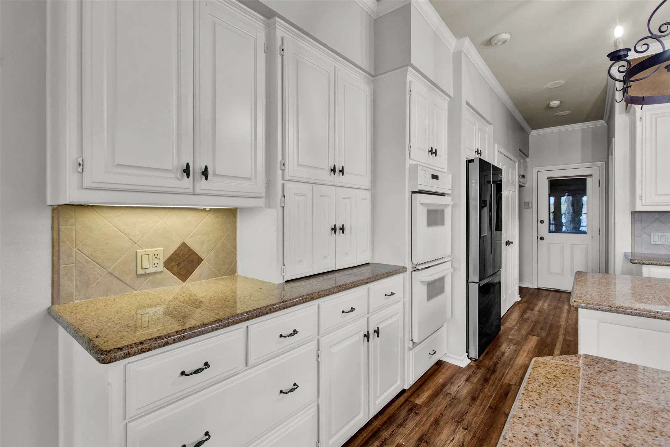White cabinetry with dark hardware, granite countertops, tile backsplash, built-in double ovens, and wood-finish flooring
