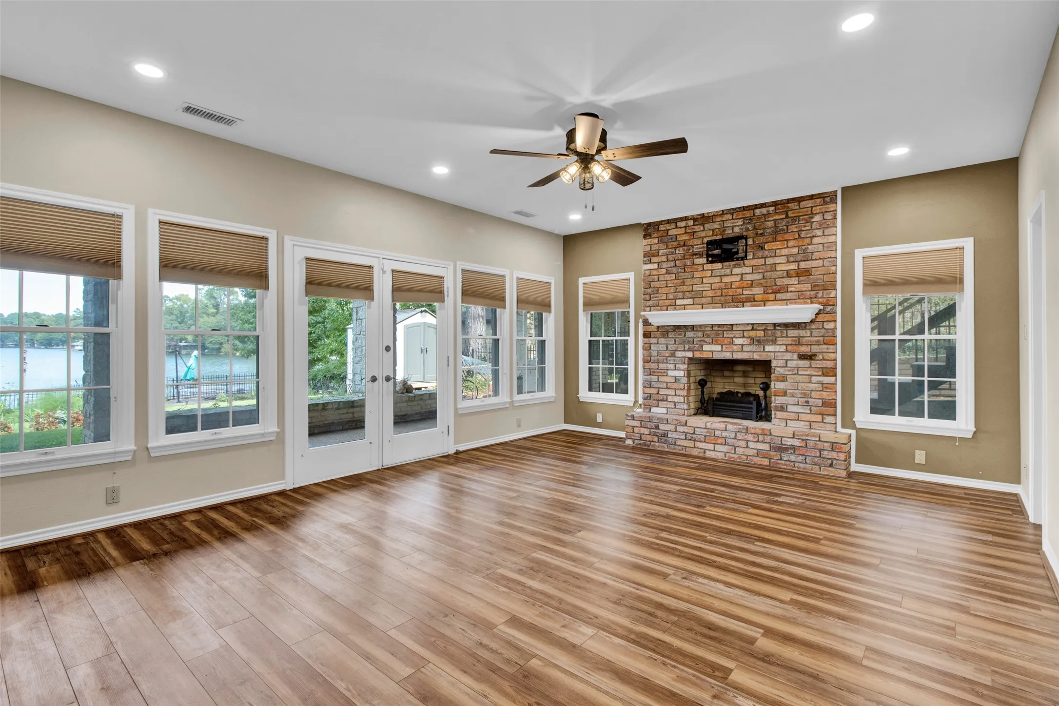 Wood-finish flooring, brick fireplace with a decorative mantel, ceiling fan, recessed lighting, and multiple windows with cellular shades
