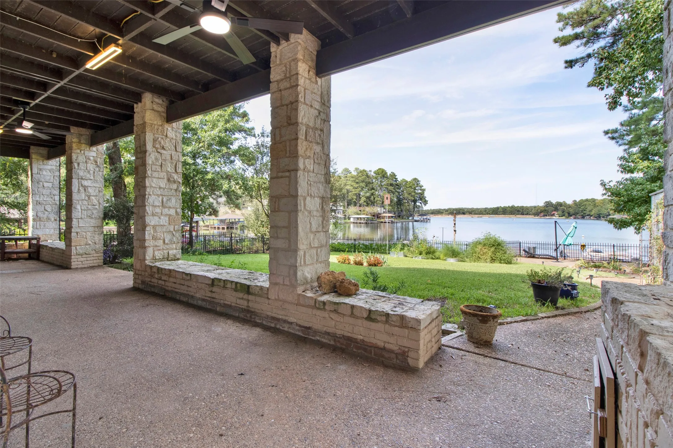 Covered patio with stone pillars and a ceiling fan, overlooking a spacious lawn and a body of water