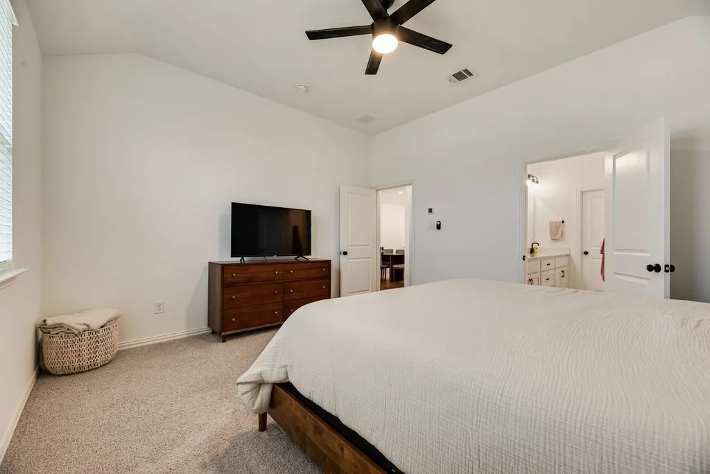 Vaulted ceiling, ceiling fan with integrated lighting, window with blinds, carpet flooring, and a door leading to a bathroom with a vanity and lighting