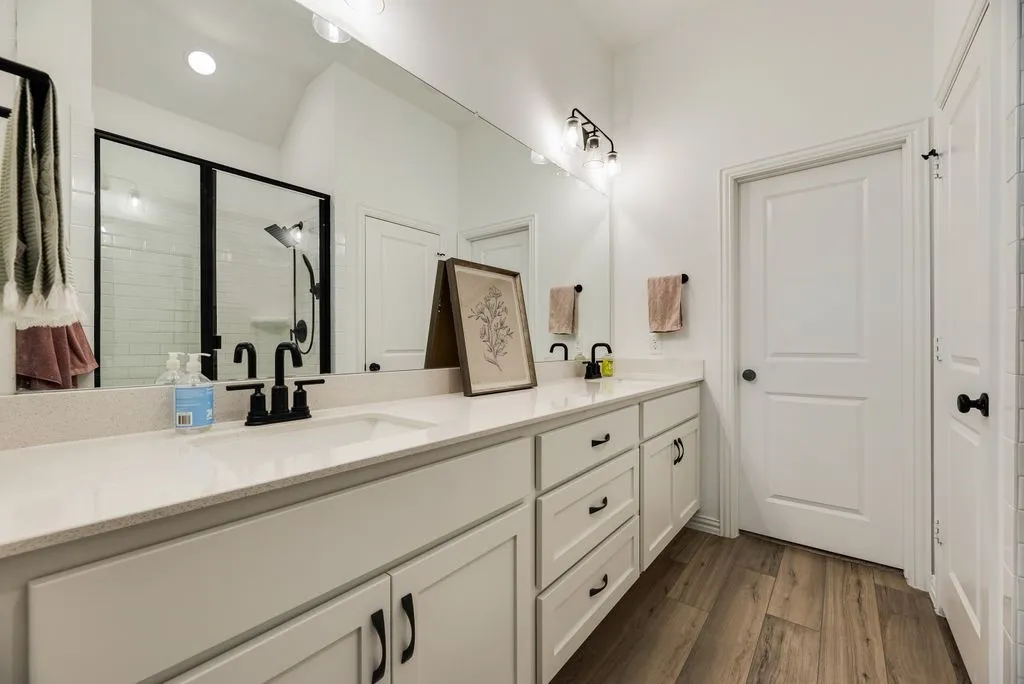 Dual vanity with light-toned countertop, matte black fixtures, extensive cabinetry, frameless mirror, and wood-finish flooring