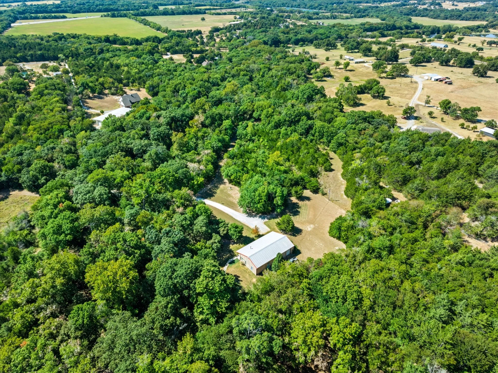 Expansive treed acreage featuring a single-story residence with a light-colored roof, a detached barn structure, and a winding gravel driveway