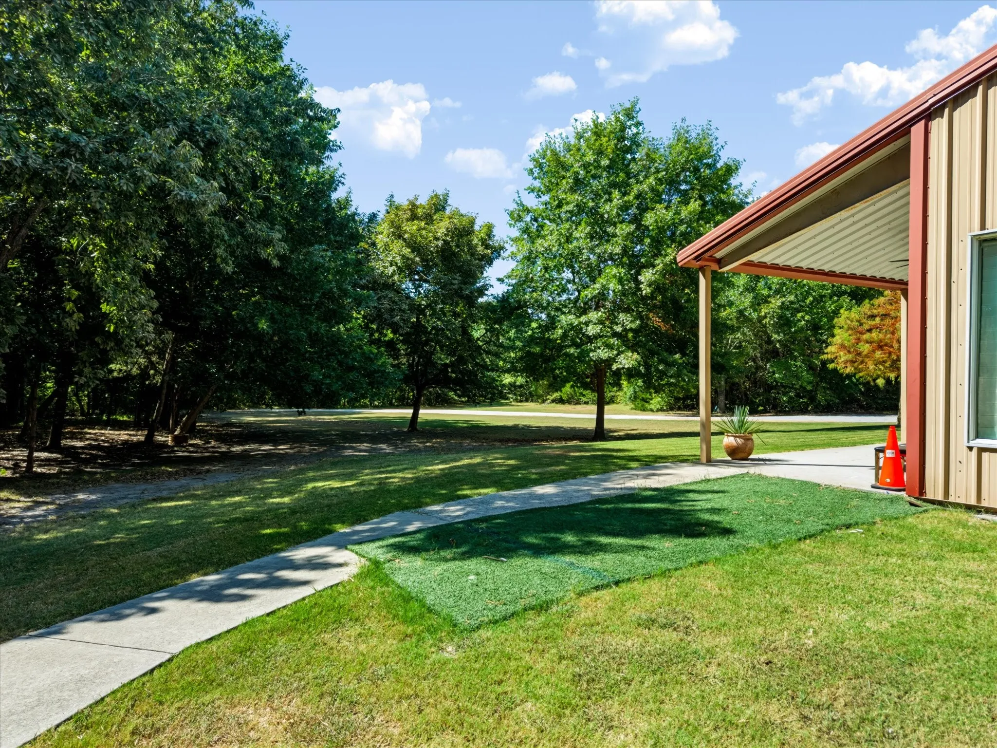 Expansive green lawn, mature trees, concrete pathway, metal siding, and a covered porch with a wood support beam