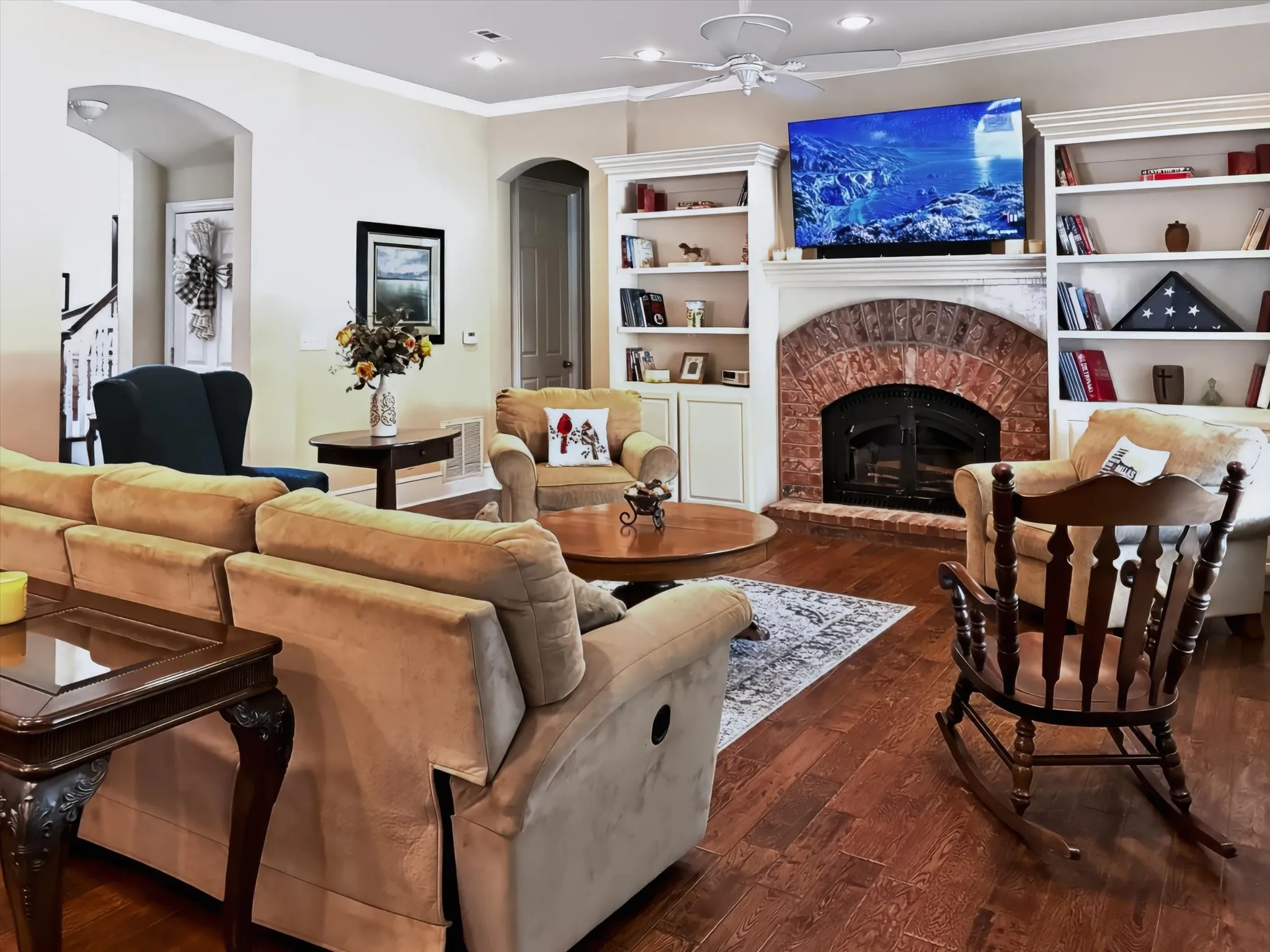 Hardwood flooring, brick fireplace with arched opening, built-in shelving, recessed lighting, and ceiling fan