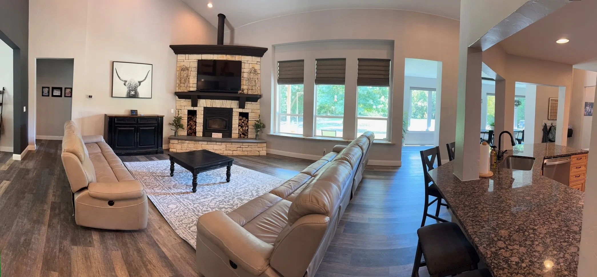 Spacious living area featuring a stone-clad fireplace with dark wood mantel, vaulted ceilings, large windows, and wood-finish flooring