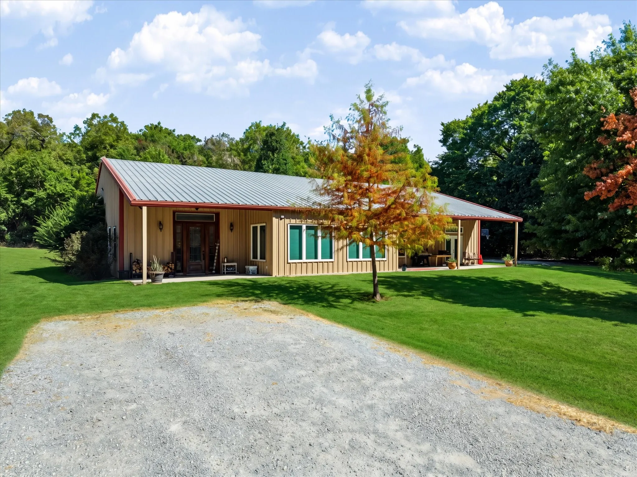 Board and batten siding with a metal roof, front porch with wood columns, gravel driveway, and surrounding mature trees