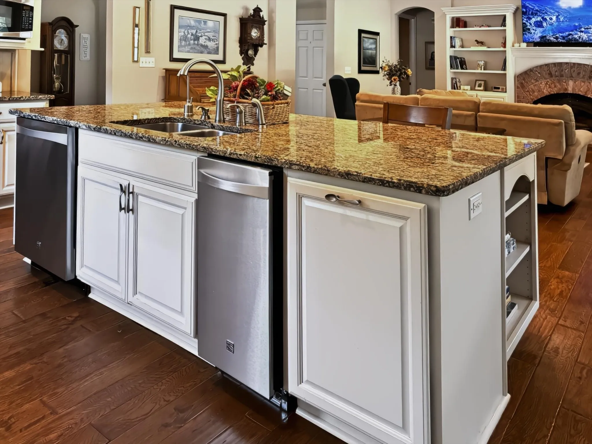 Kitchen island featuring a dual-basin stainless steel sink, granite countertop, built-in dishwasher, white cabinetry, and wood-finish flooring