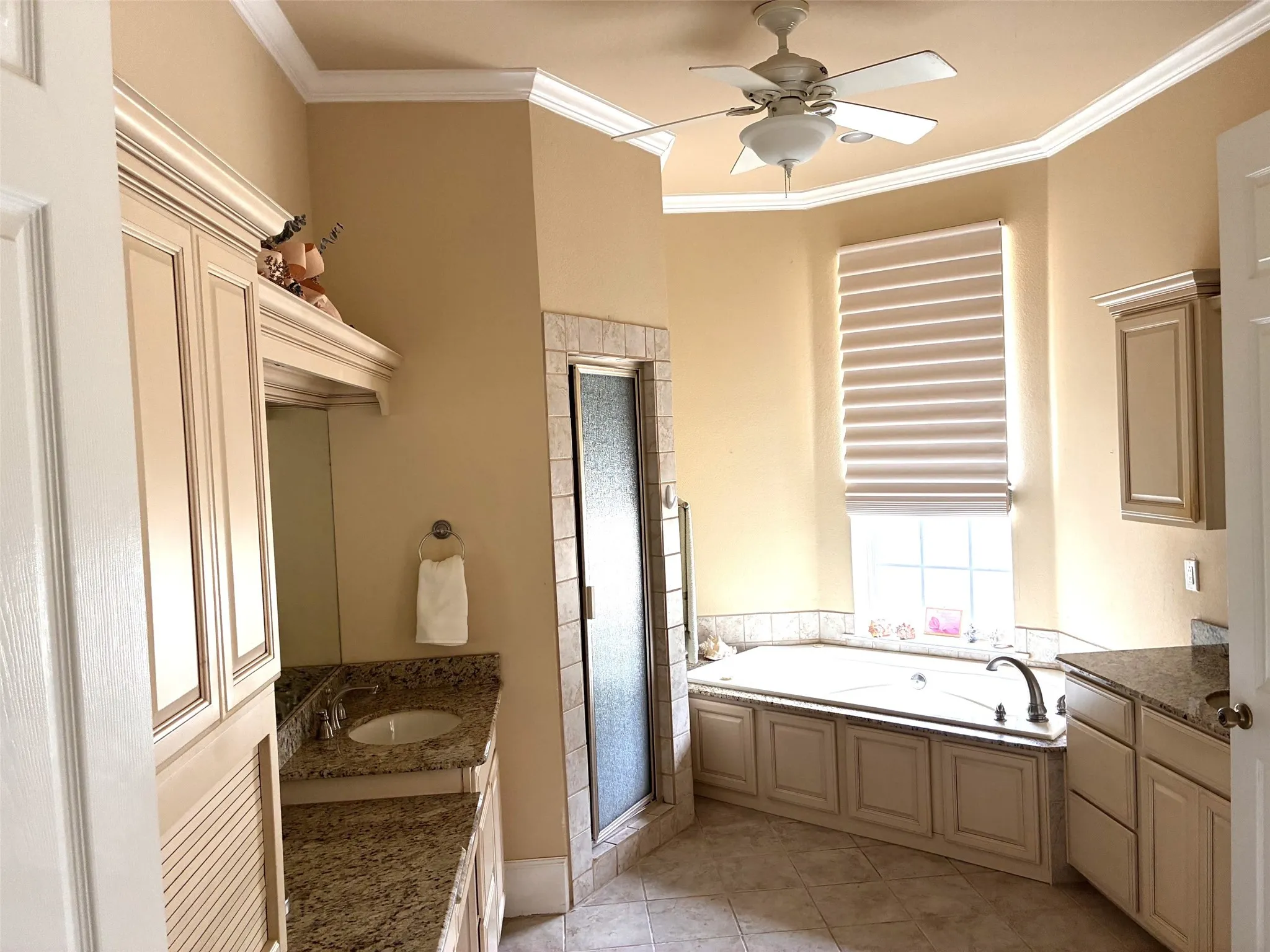 Dual vanities with granite countertops, a built-in soaking tub, a stand-alone shower with a glass enclosure, a ceiling fan, and crown molding
