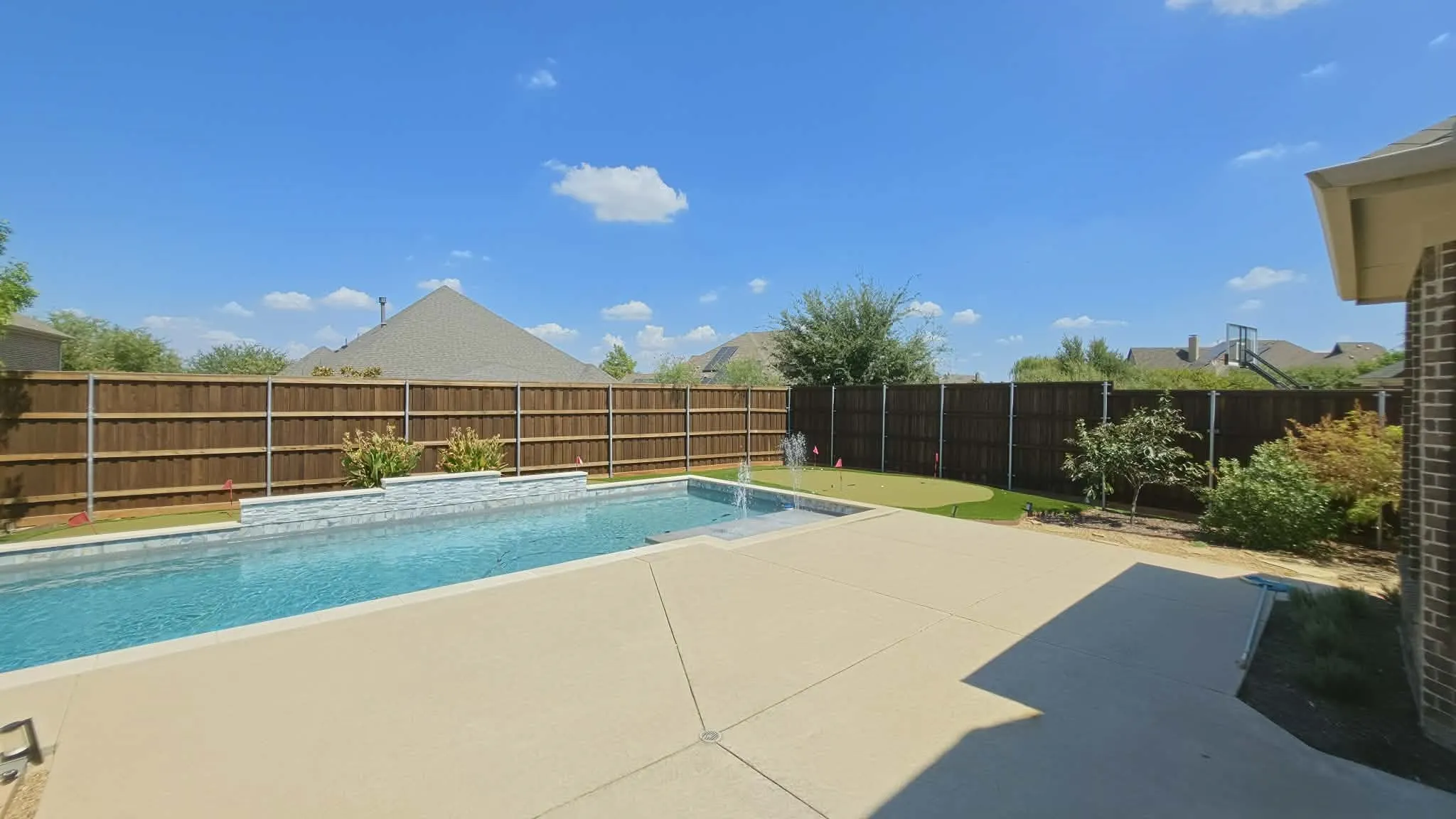 Backyard featuring a rectangular swimming pool with integrated water features, concrete decking, a putting green, and a privacy fence