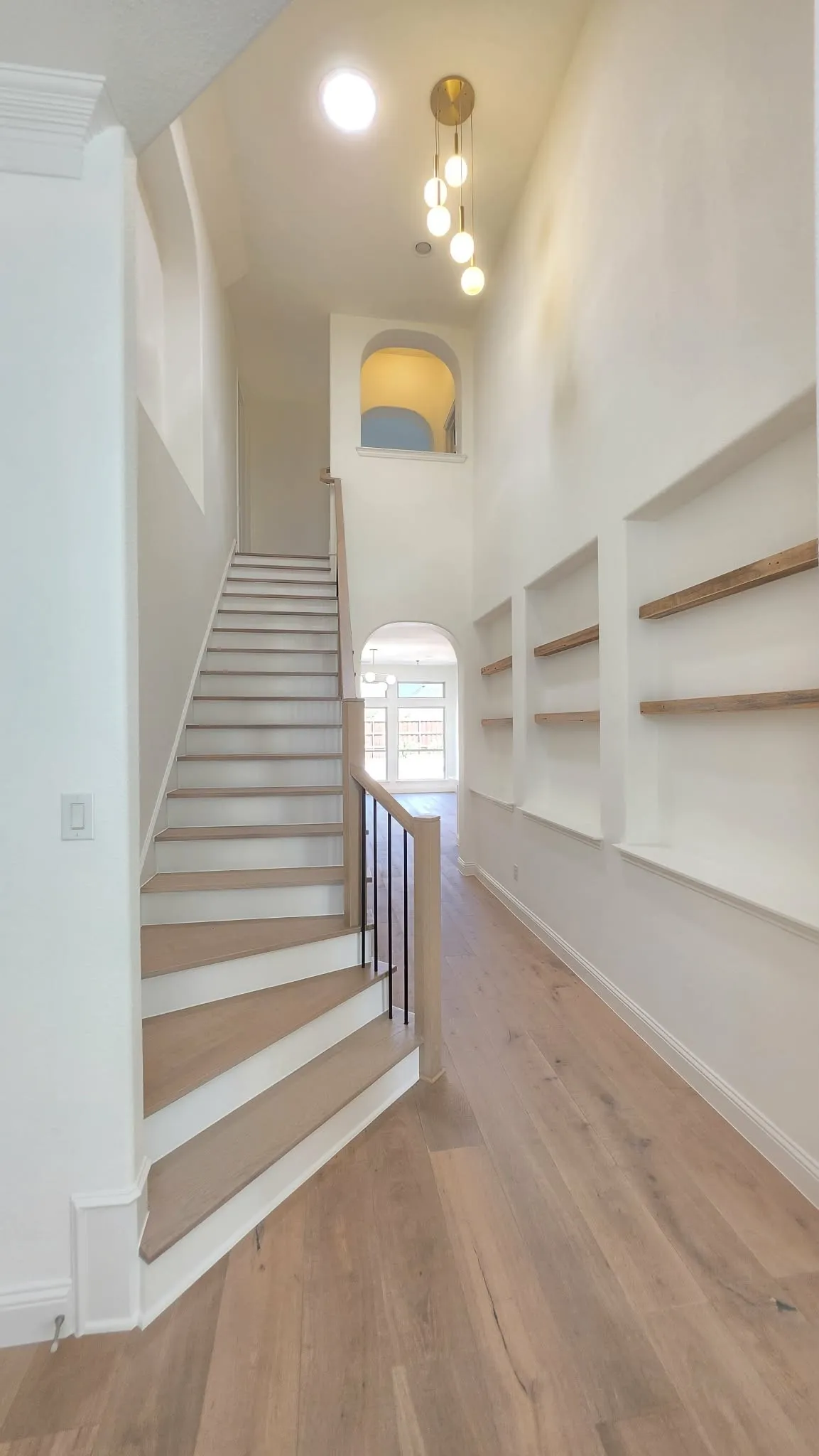 Hardwood-style flooring throughout, prominent staircase with wood-finish handrail, built-in wall shelving, arched wall features, and a modern ceiling light fixture