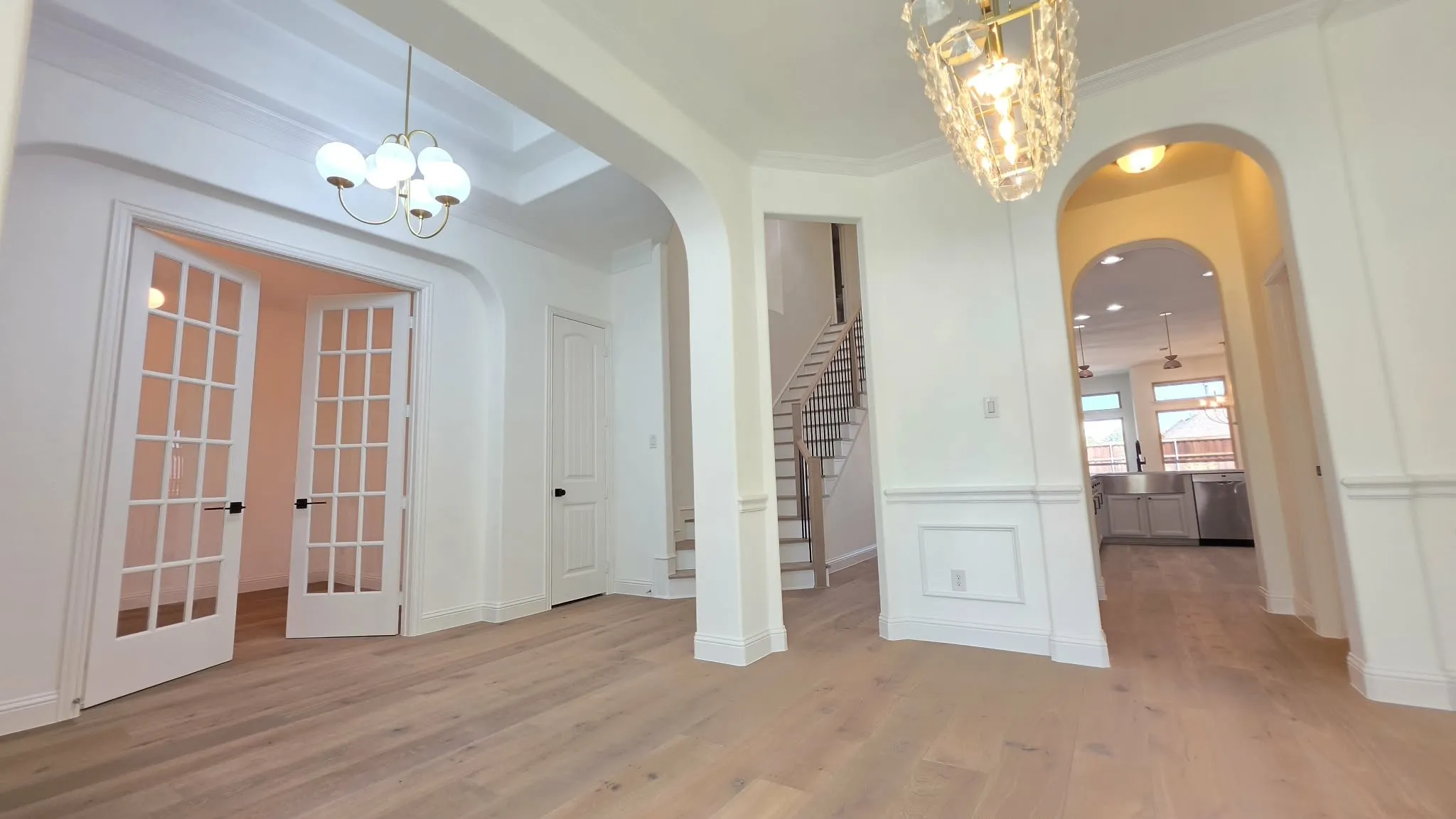 Light wood-finish flooring, two decorative chandeliers, arched doorways, French doors, and wainscoting