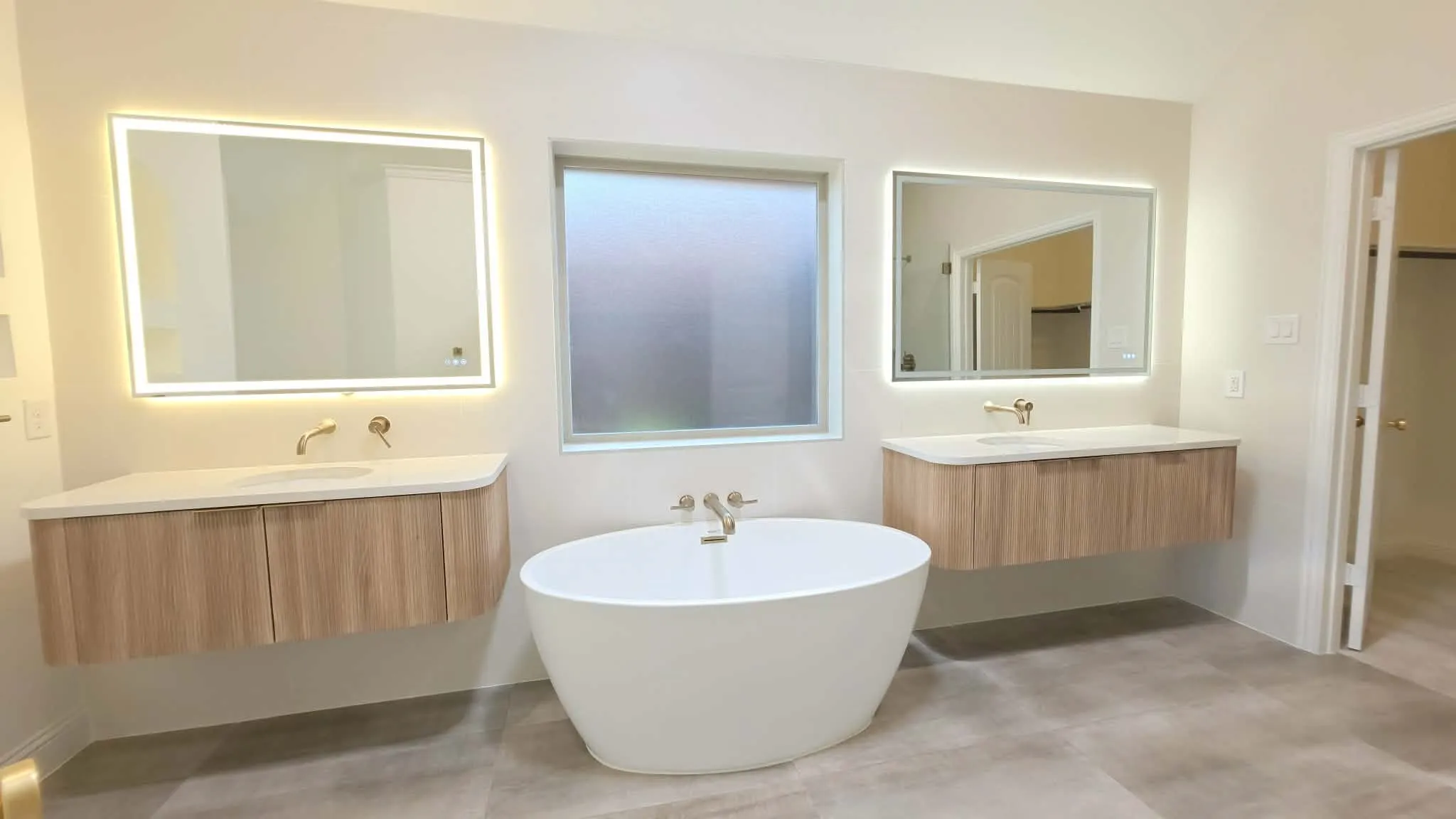 Dual floating vanities with light wood-finish cabinetry, illuminated mirrors, and a freestanding soaking tub