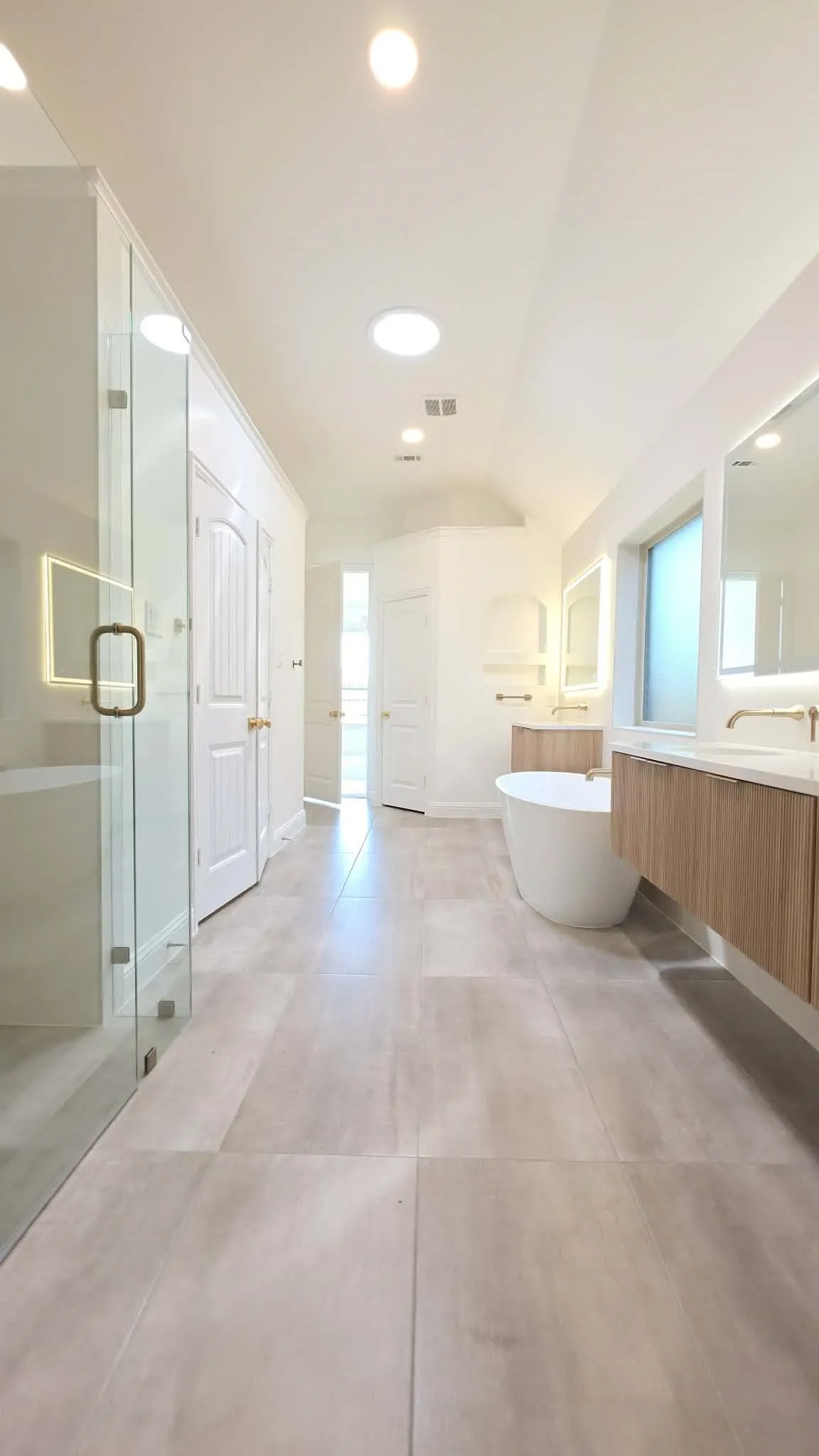 Spacious bathroom featuring large format tile flooring, a glass-enclosed shower, dual floating wood-finish vanities, and a freestanding soaking tub