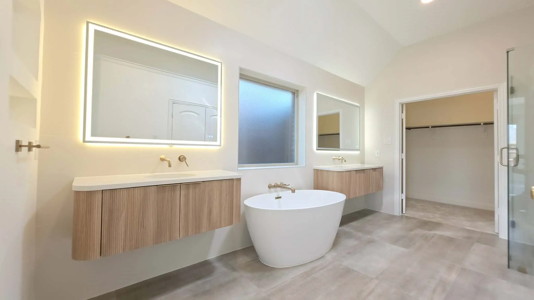 Vaulted ceiling with an illuminated mirror, wood-finish floating vanity, freestanding soaking tub, and a glass-enclosed shower