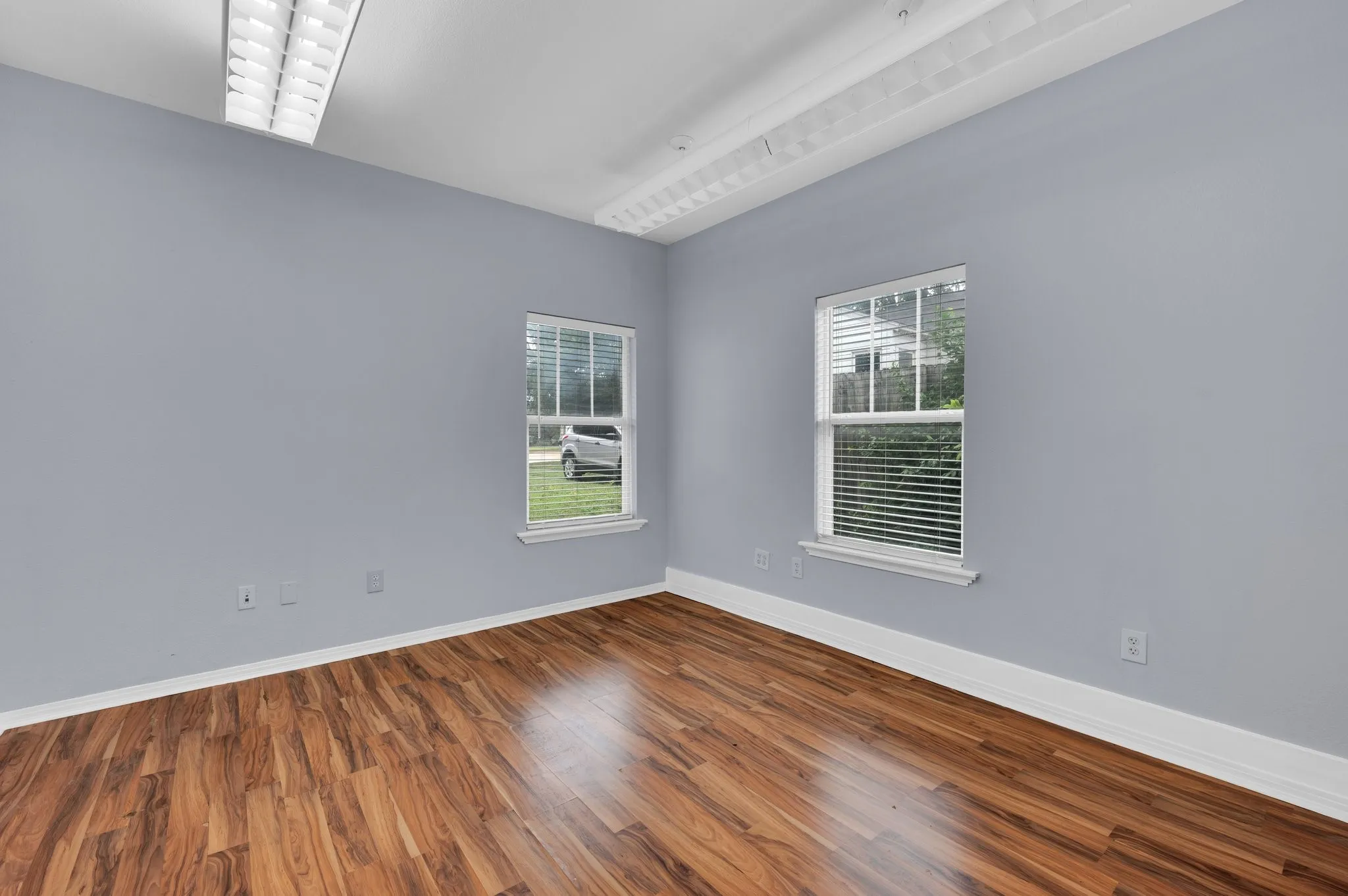Room featuring wood-finish flooring, two windows with horizontal blinds, light gray wall paint, white baseboards, and overhead fluorescent lighting